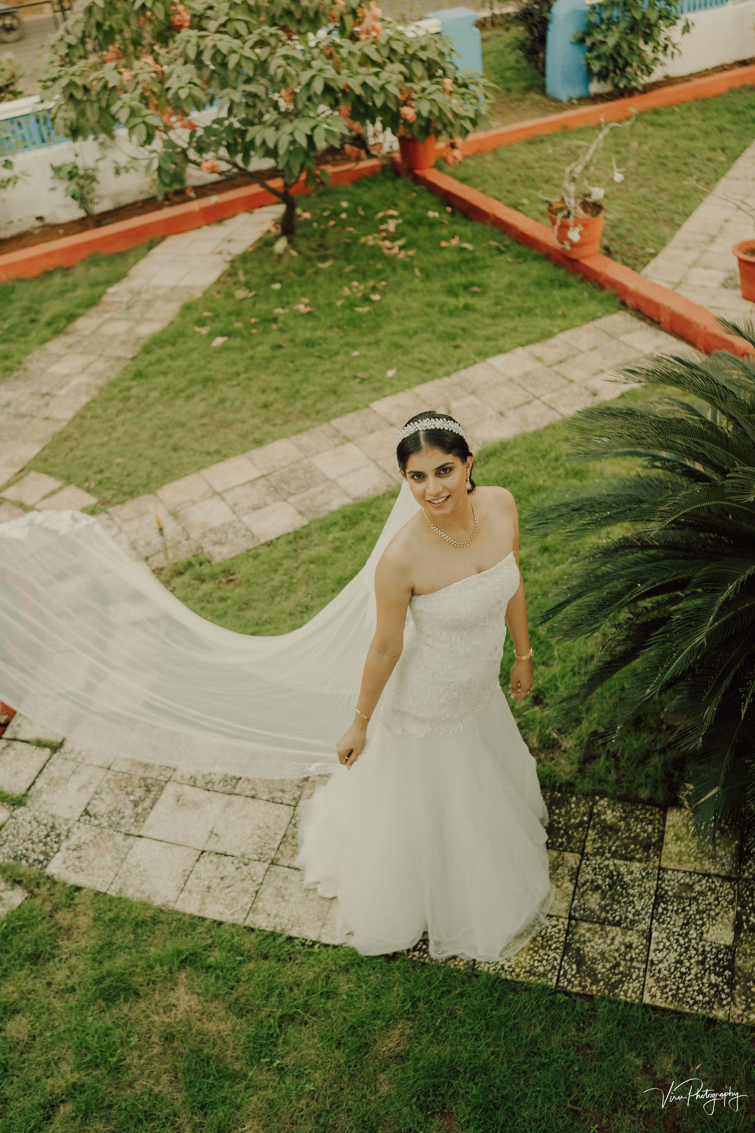 A person in a flowing white strapless wedding gown stands on stone steps surrounded by lush garden landscaping.