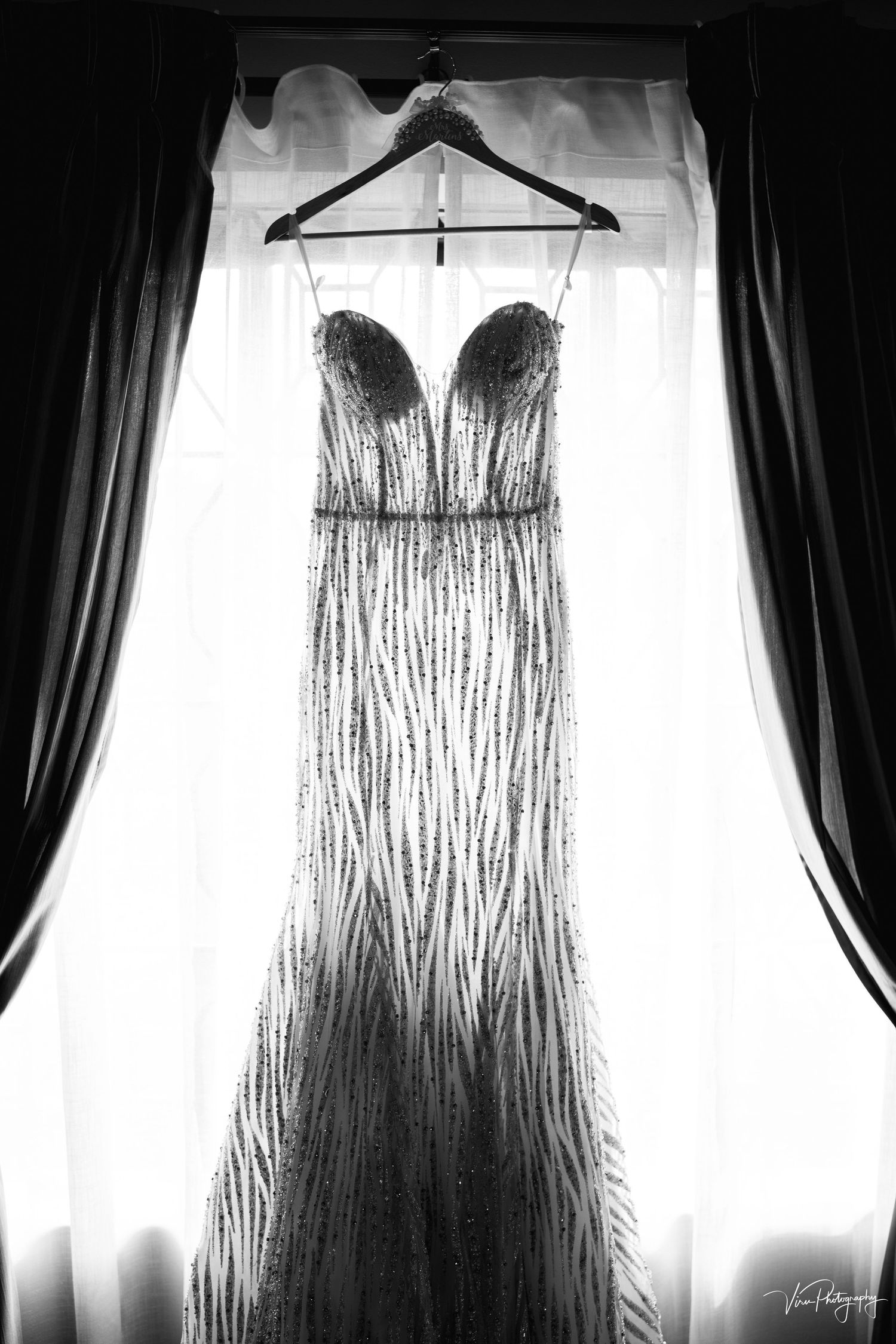 A black and white photograph of a strapless evening gown with dramatic beading hanging in front of a window with curtains.