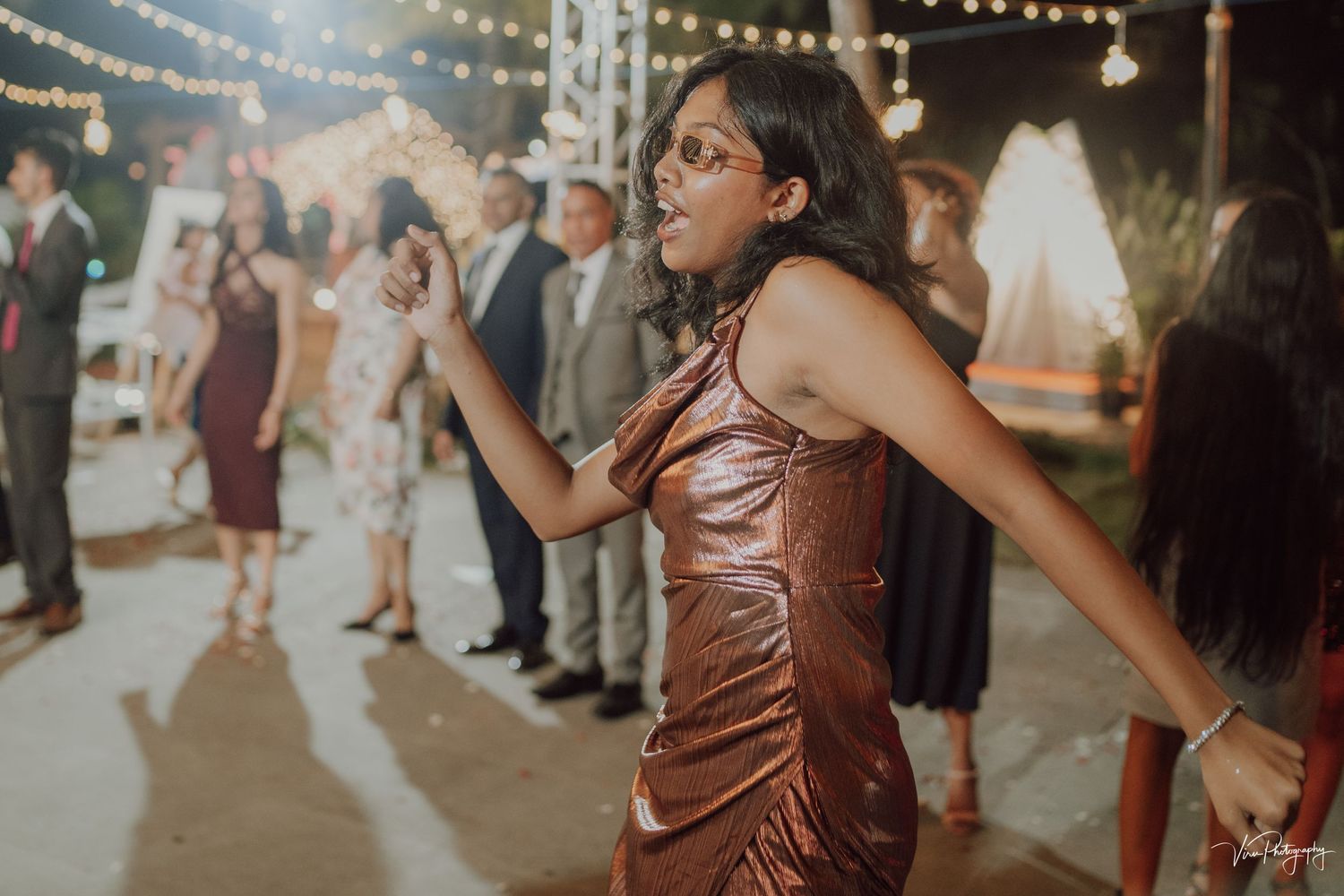 Person in shimmering brown dress dances energetically at an outdoor celebration with string lights overhead.