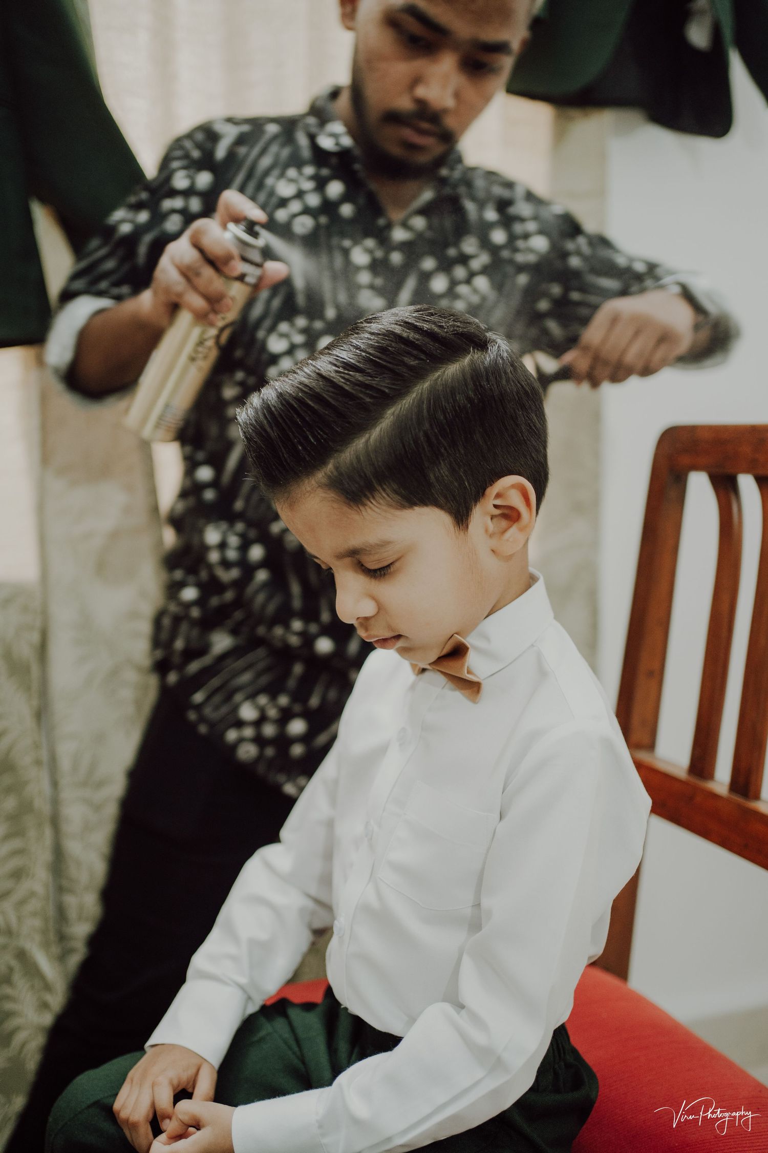 A child sits in a white dress shirt while someone styles their neat dark hair with professional grooming tools.