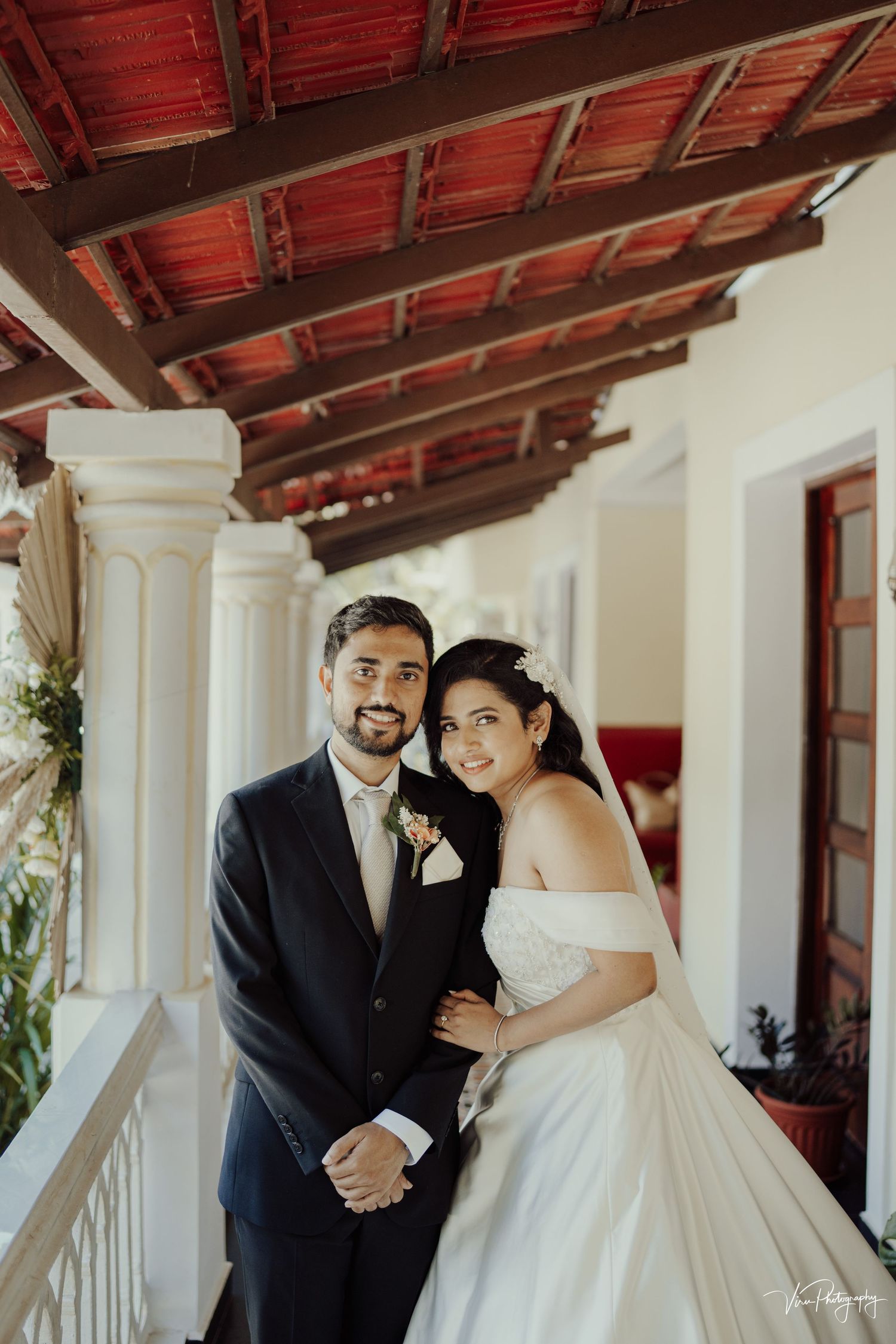 A couple stands together under a covered Spanish-style porch with white columns and terracotta roof tiles.