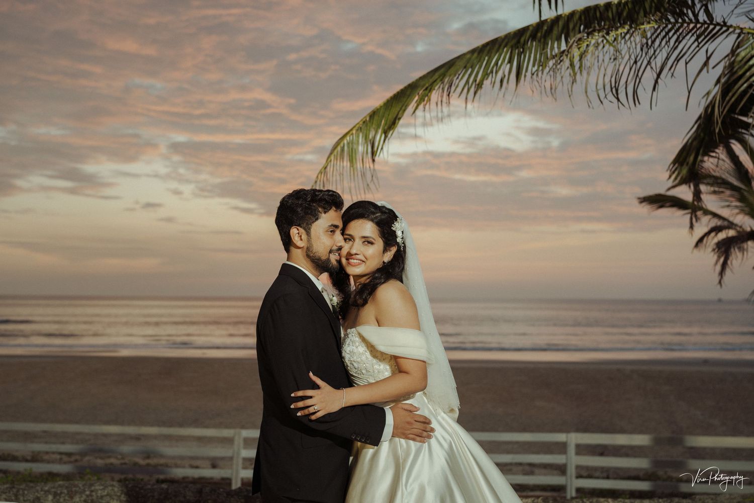 A bride and groom embrace on a beach at sunset with palm trees and dramatic clouds in the background.