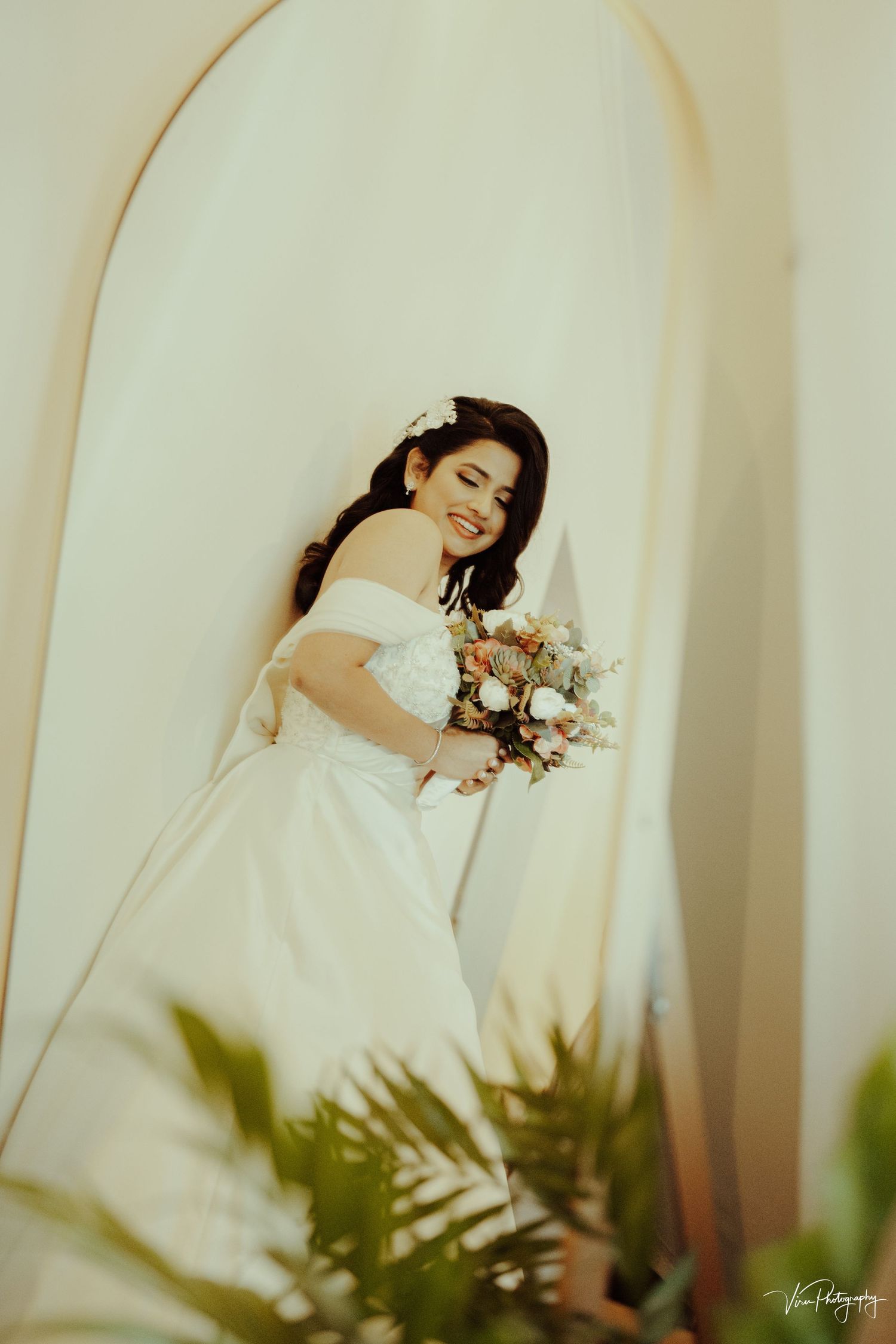 Vintage-style bridal portrait showing an off-shoulder white wedding gown and pastel bouquet through an arched doorway.