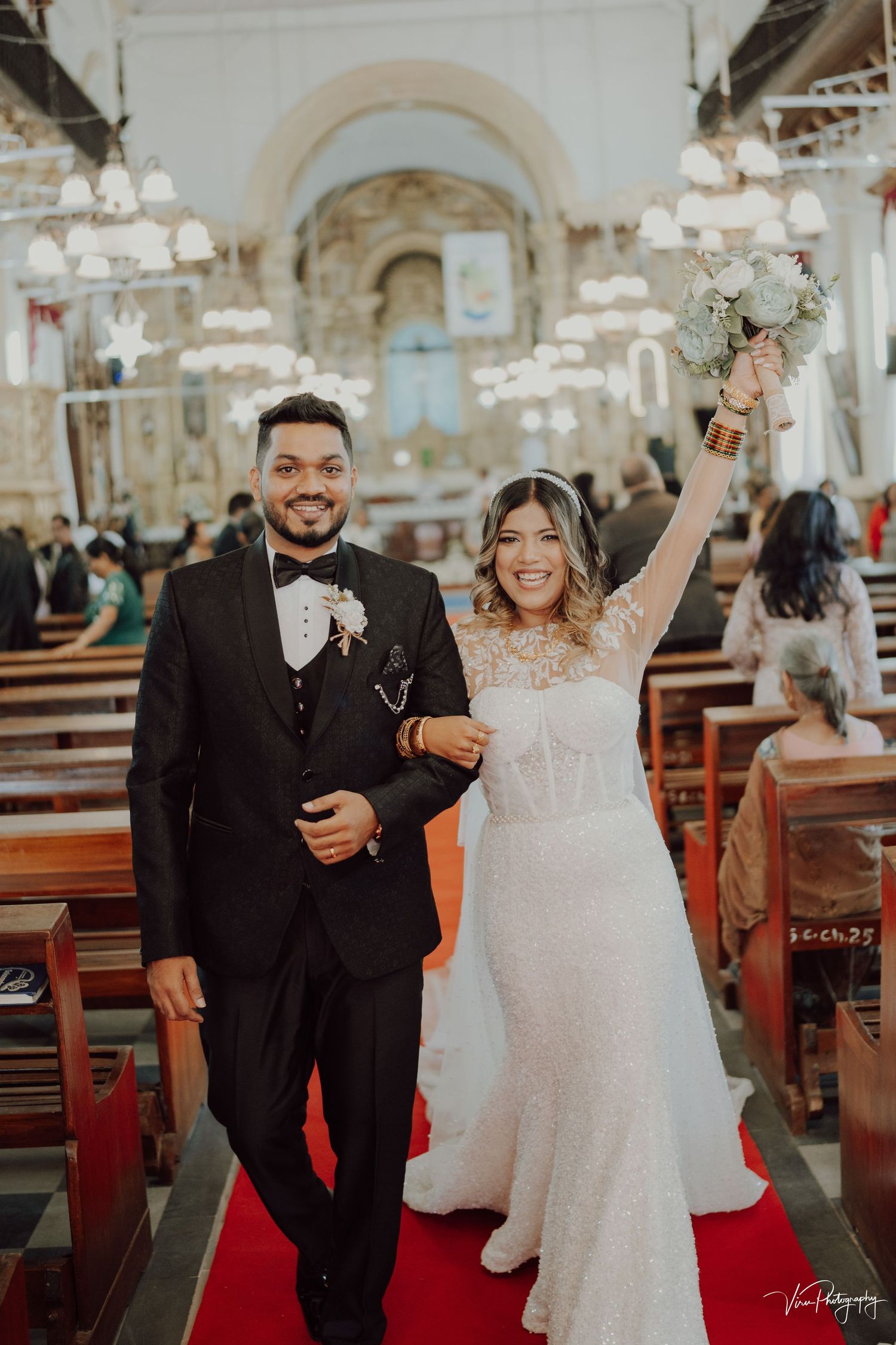 A joyous wedding couple walks down a church aisle decorated with chandeliers and wooden pews.