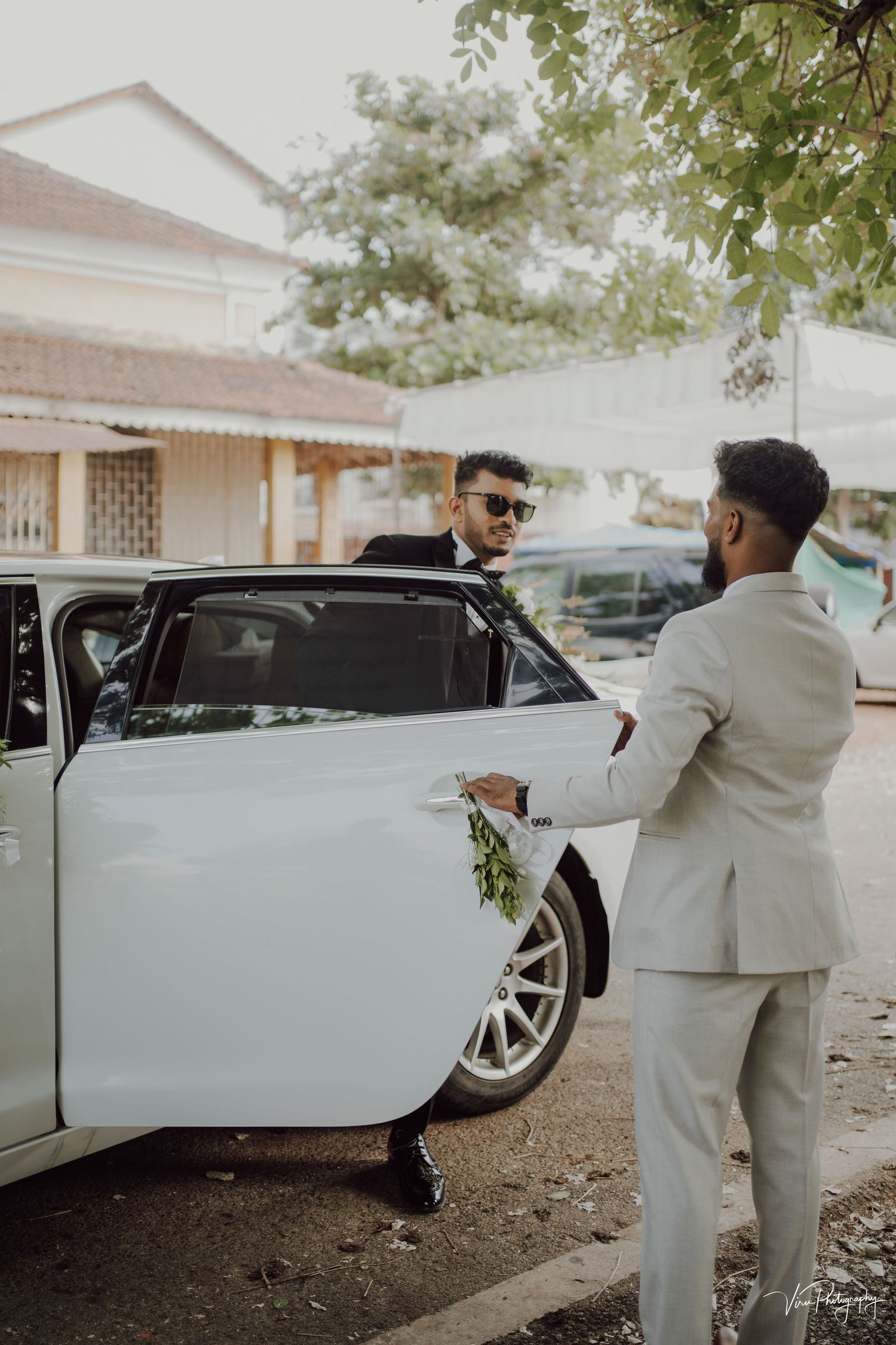 Two people in formal attire interact near a white luxury car parked outside a building.
