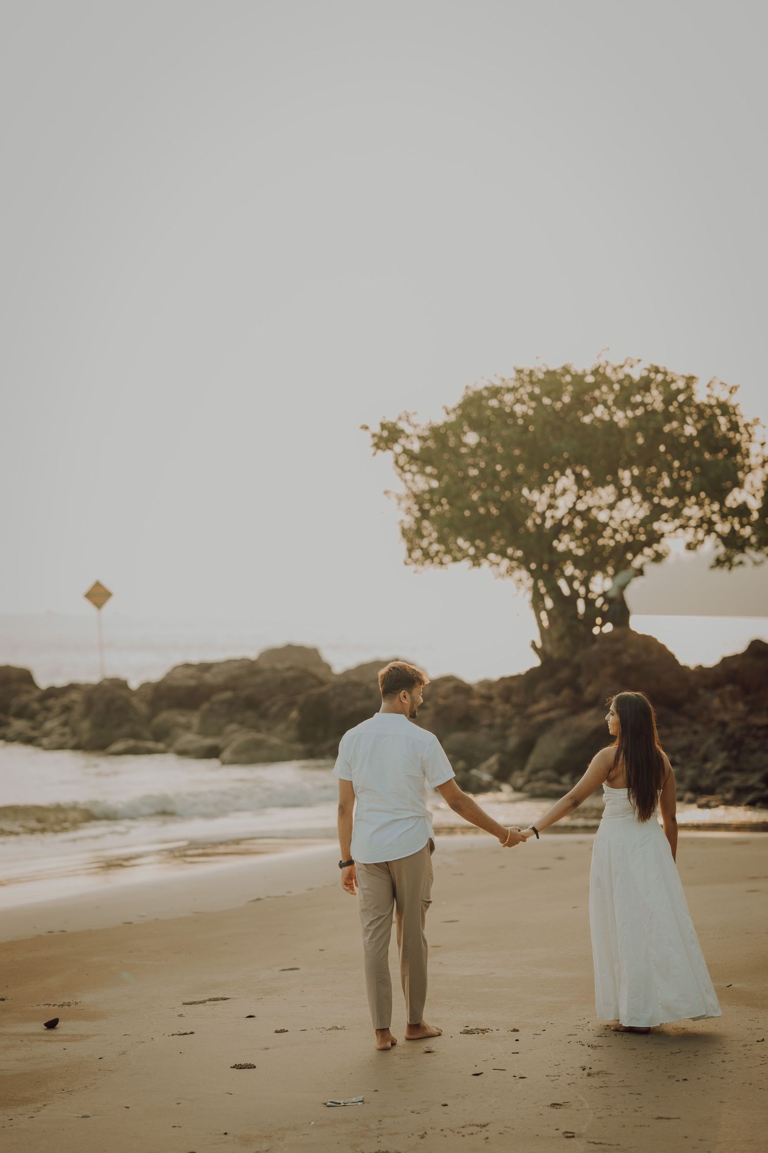 A couple walks along a scenic beach at sunset holding hands while wearing light colored formal attire.