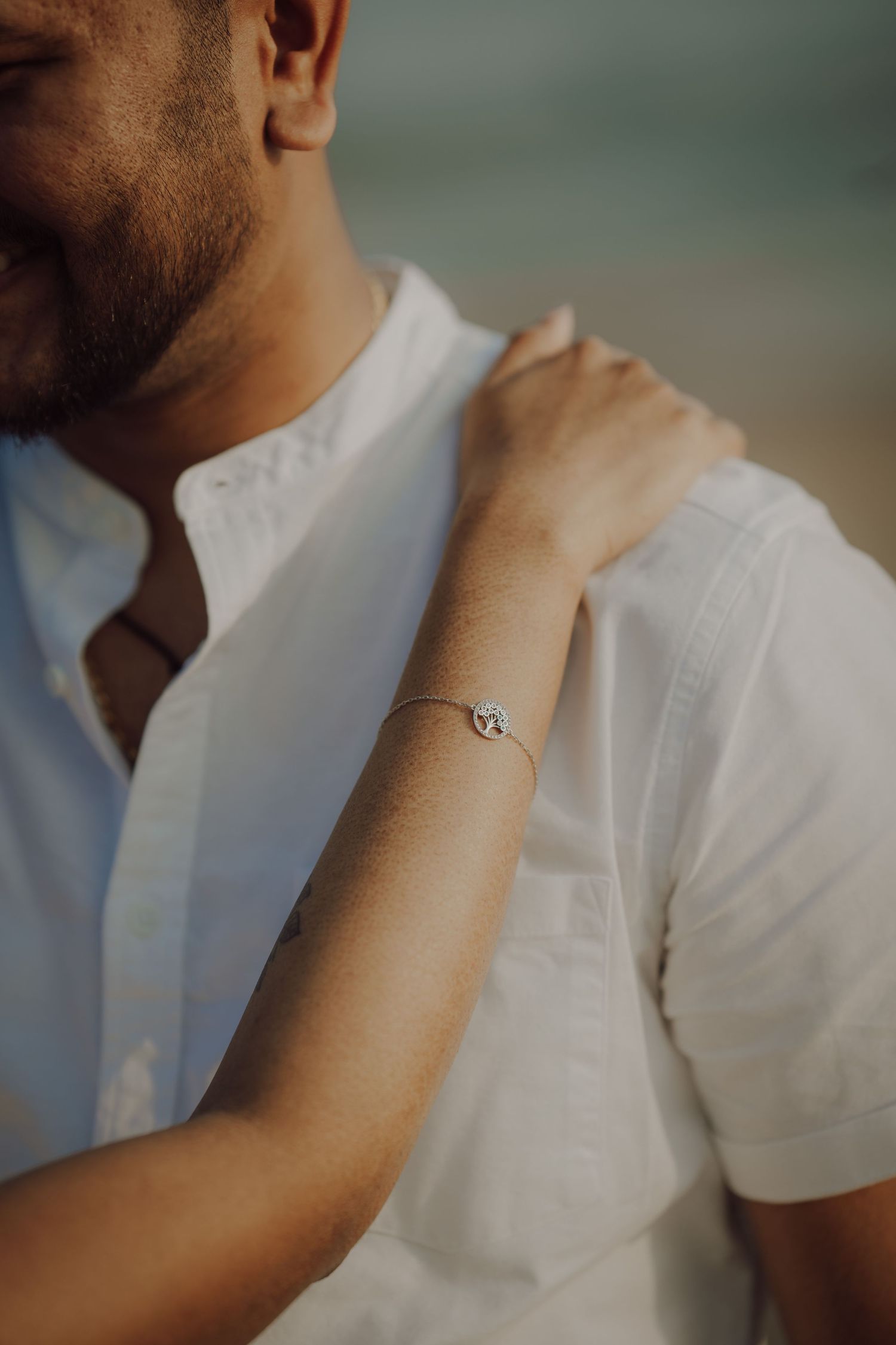 Close-up shot of a delicate silver bracelet on an arm against a white button-up shirt in soft lighting.