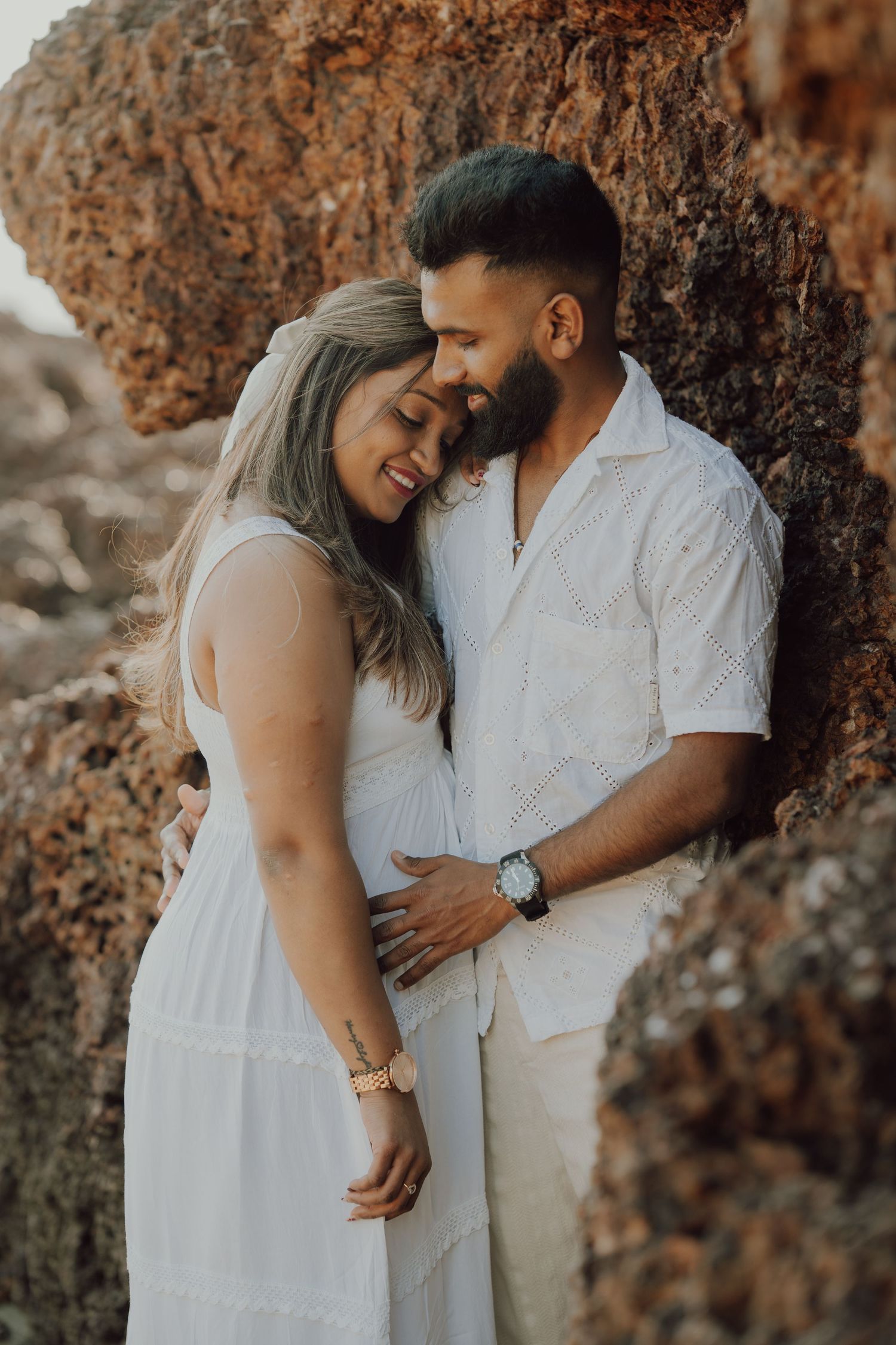 A romantic couple in white outfits share tender moments against a rocky cliff backdrop during a photoshoot.