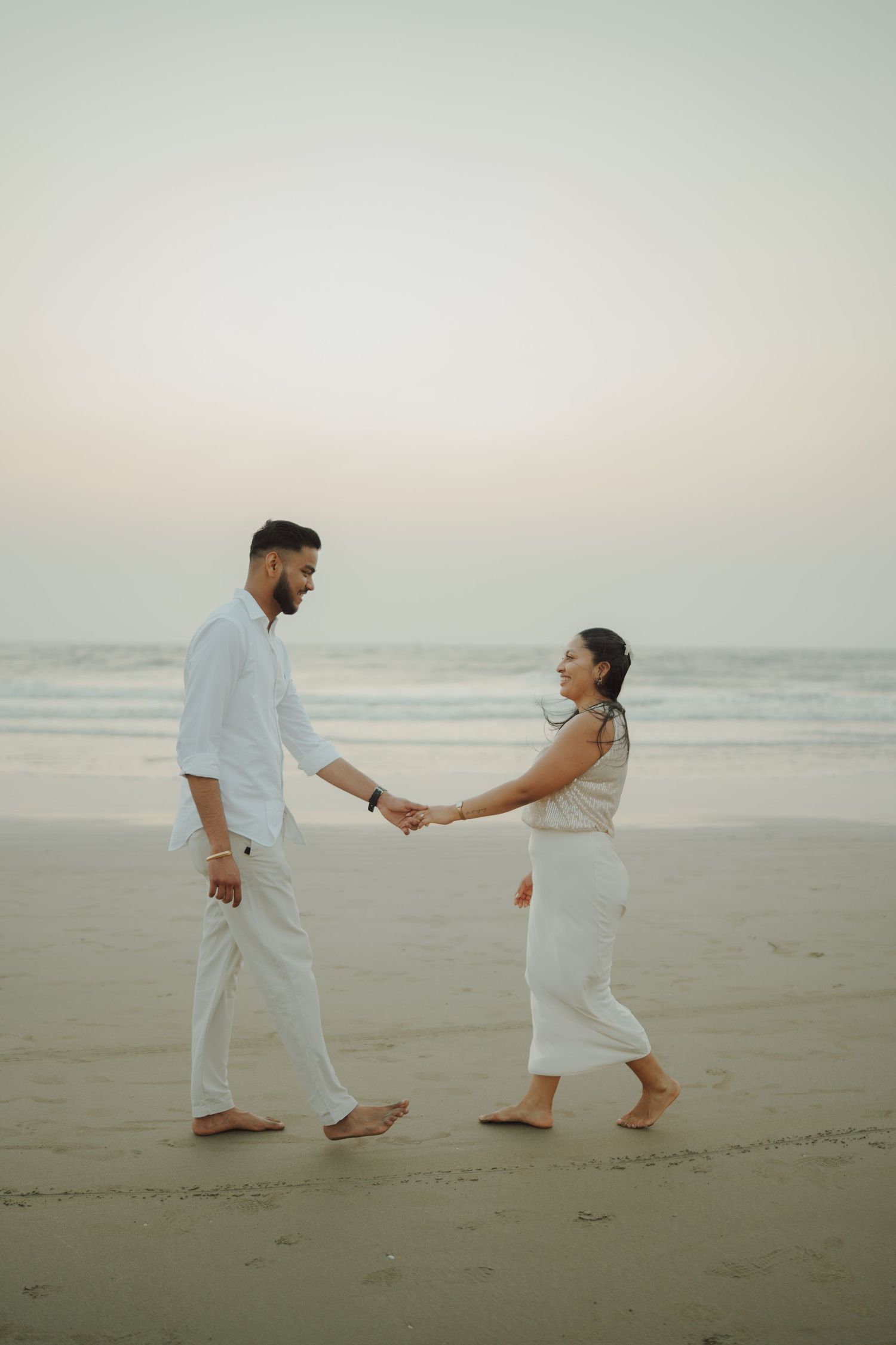 Romantic couple holding hands on a serene beach at dusk, both wearing white outfits.