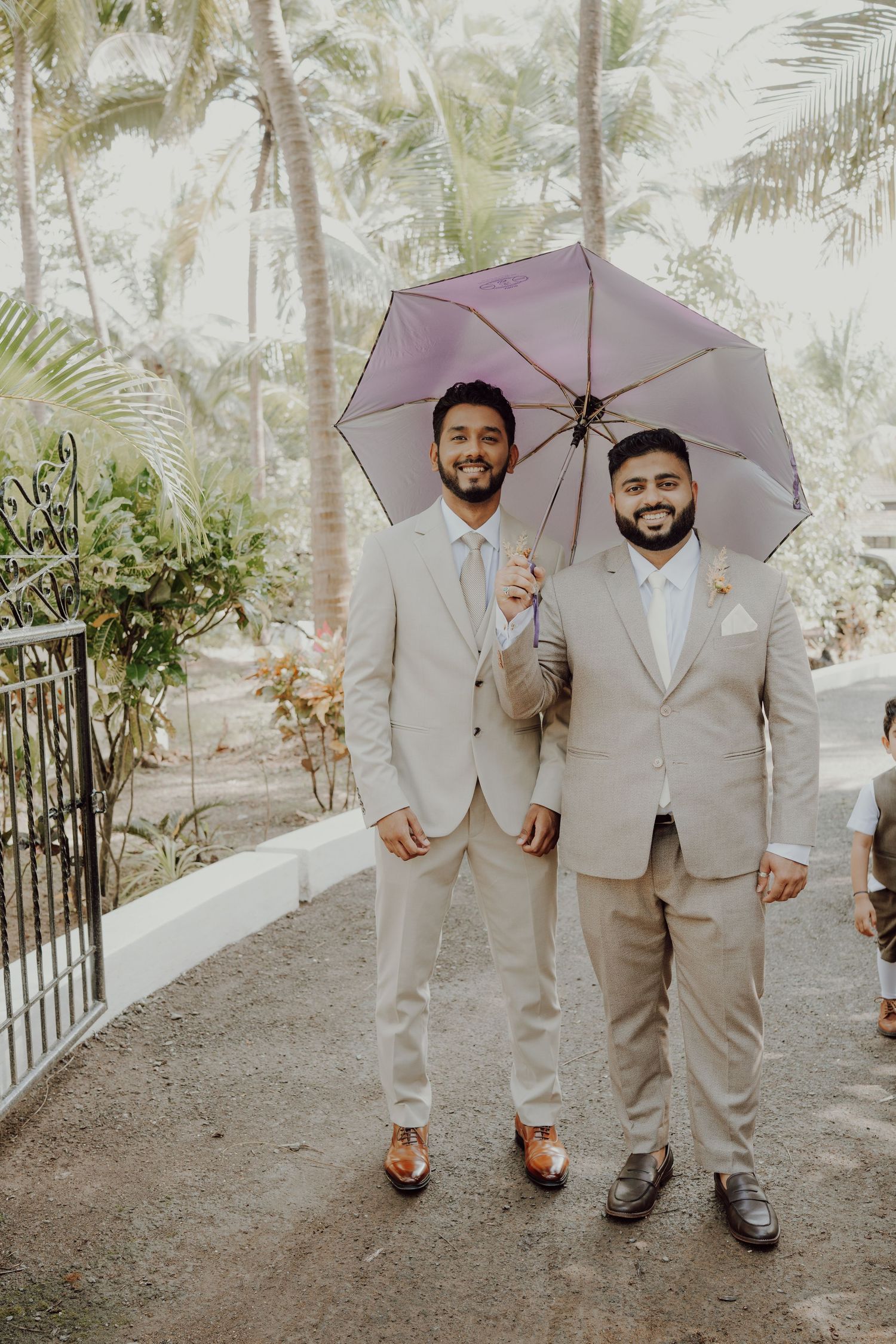 Two smiling grooms in matching beige suits share a purple umbrella on a tropical garden path surrounded by palm trees.