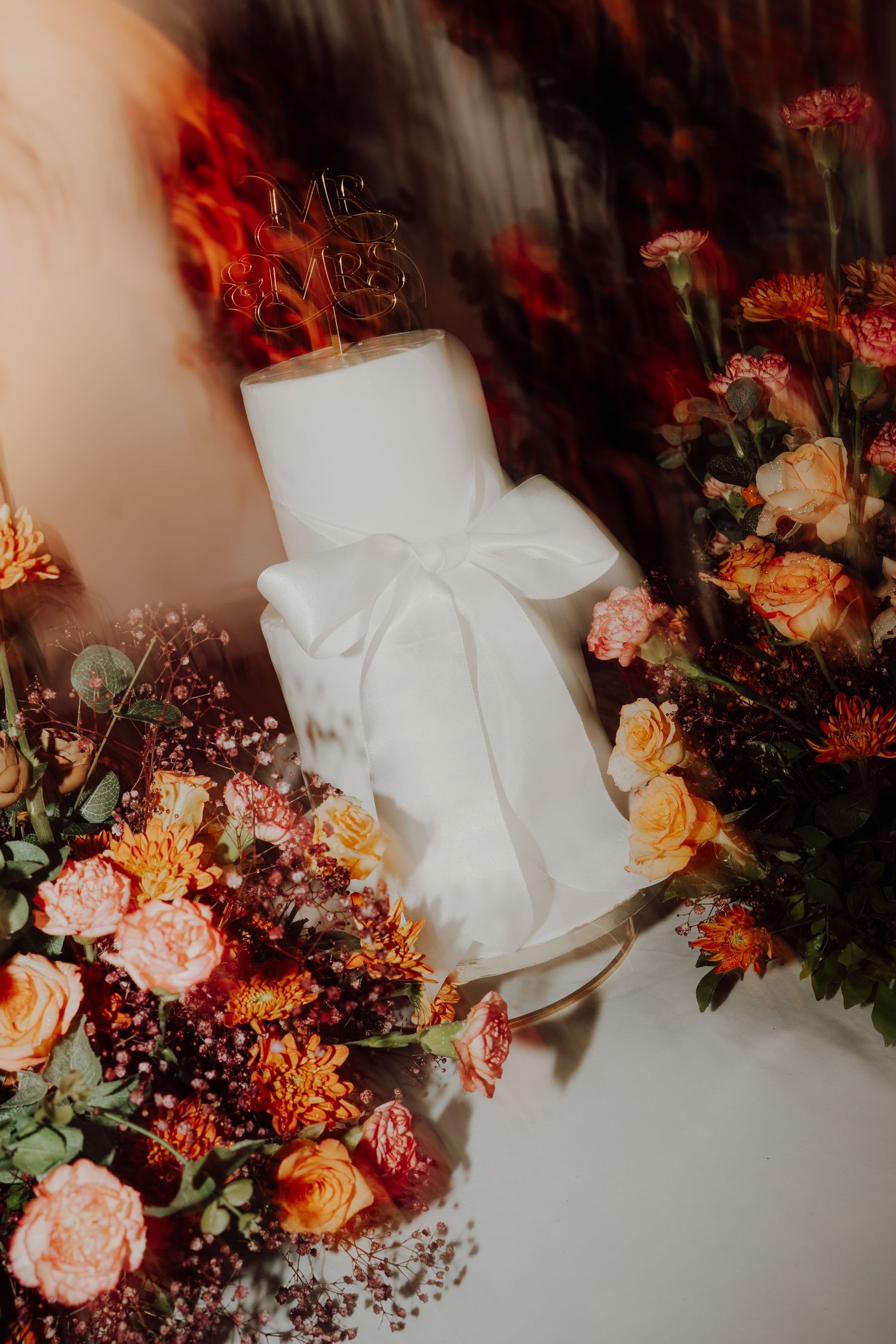 White wedding cake tied with a bow sits among orange and pink roses and dried flowers in a dreamy, blurred shot.