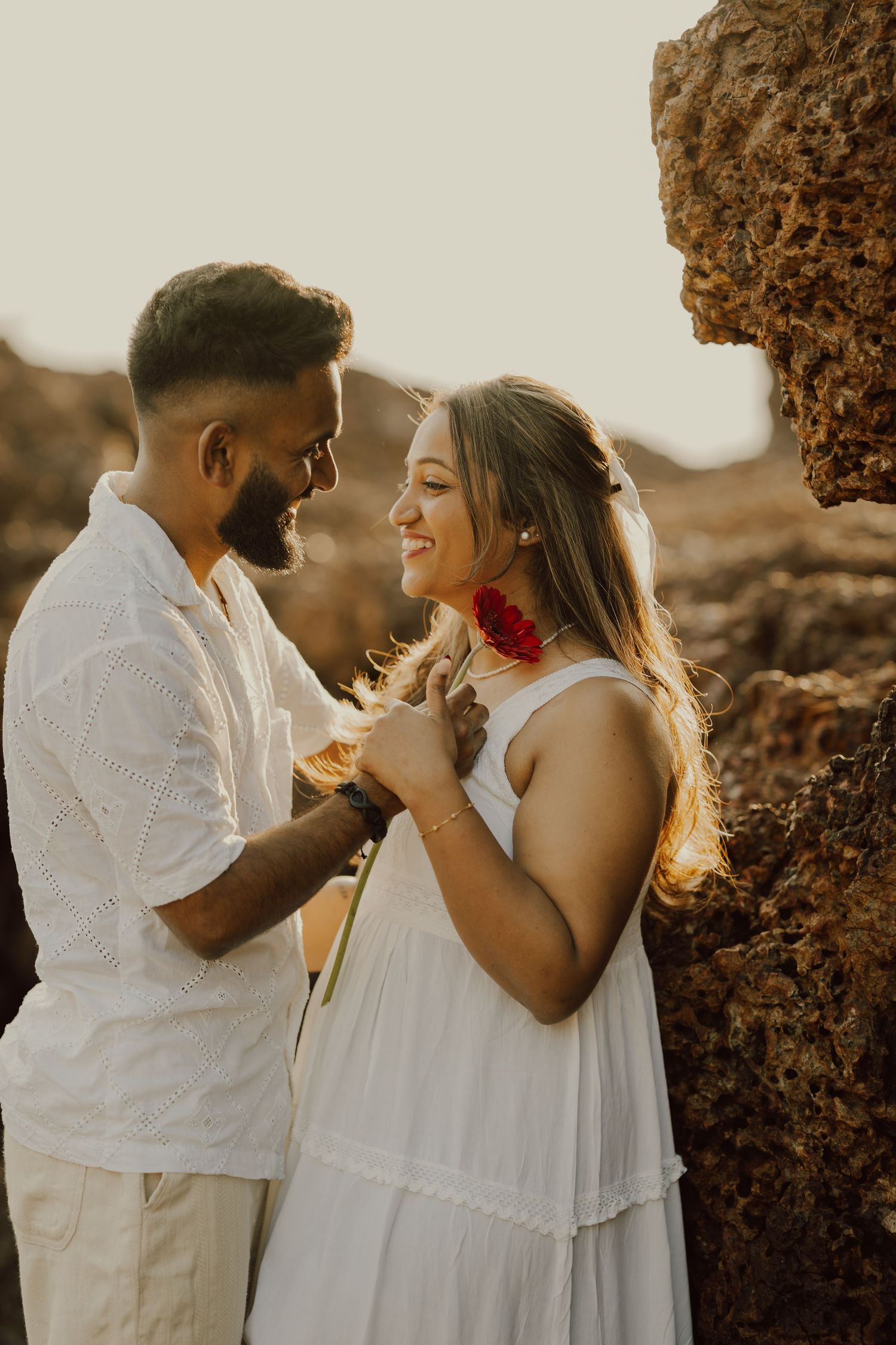 A couple shares intimate moments in white outfits against a warm sunset backdrop at a stone wall location.