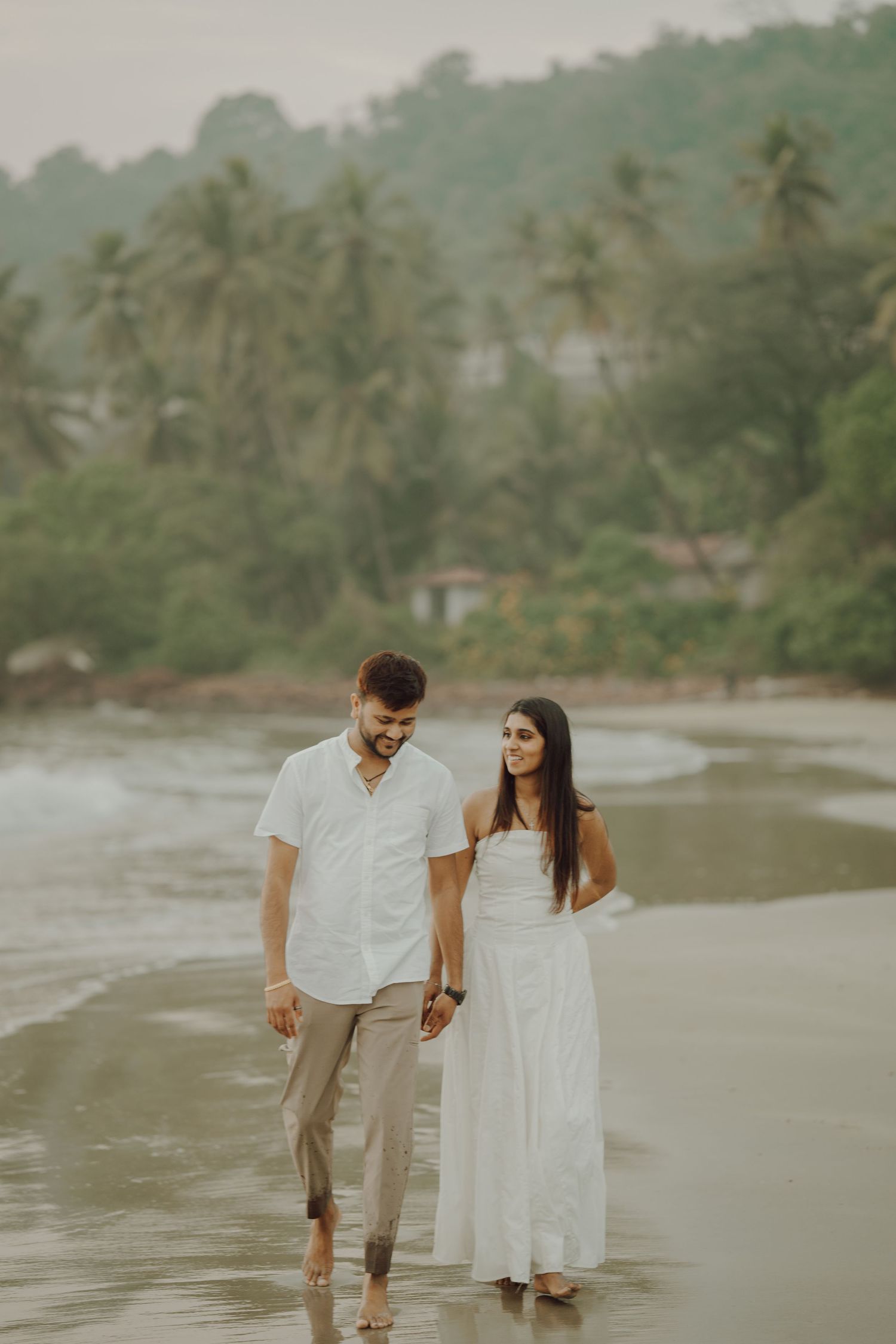 A couple in white and beige outfits walks together along a misty tropical beach with palm trees in the background.