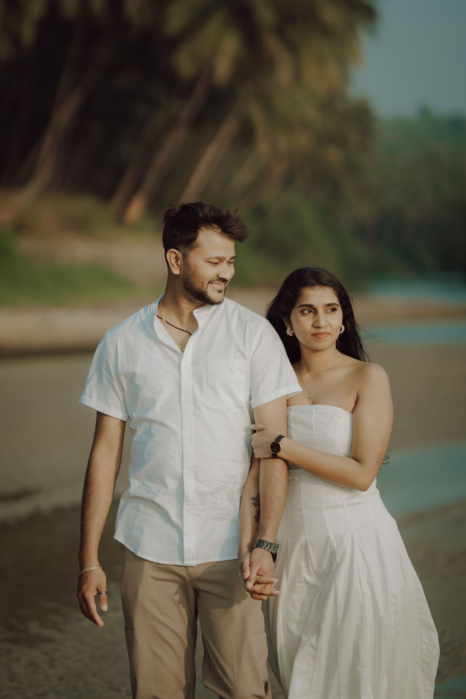 A romantic beach photoshoot with a couple walking along the shore in white and beige outfits at sunset.