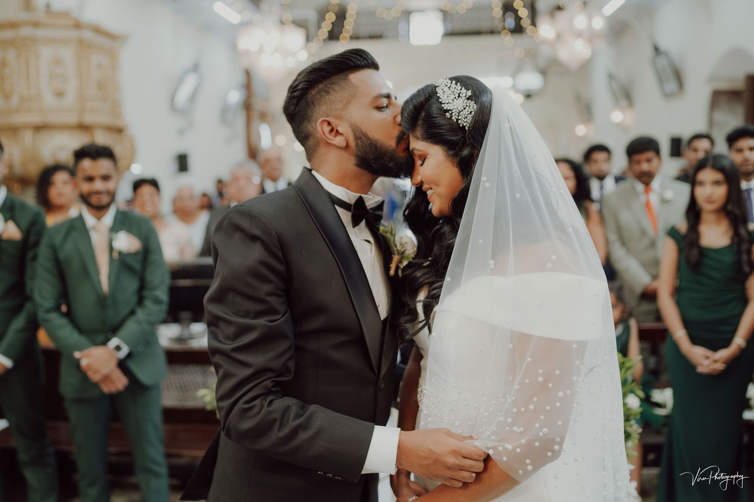 Newlyweds share an intimate moment during their wedding ceremony in church with guests in green attire looking on.