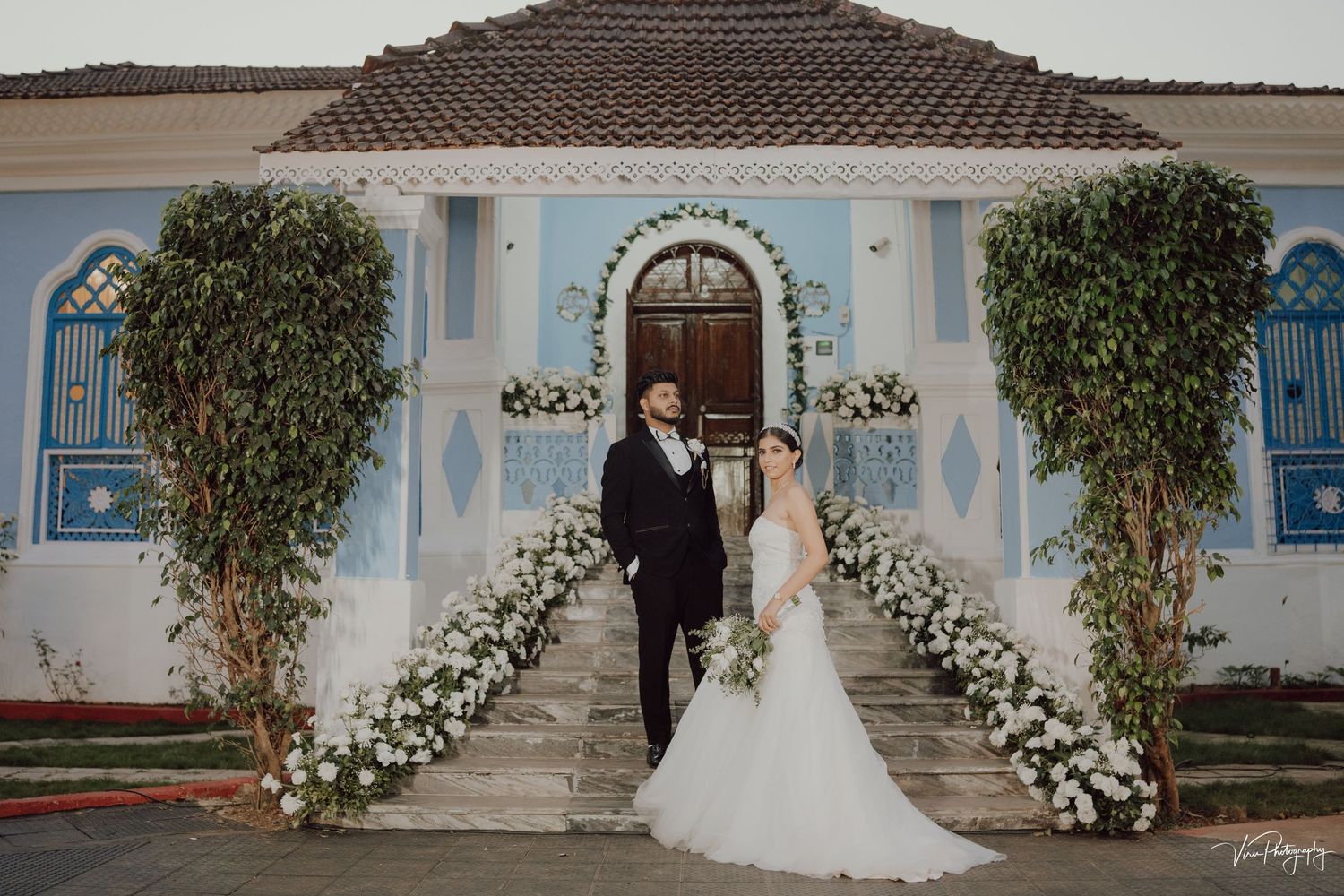 A couple stands at the entrance of a Spanish-style villa with white floral arrangements lining the walkway and blue accents.
