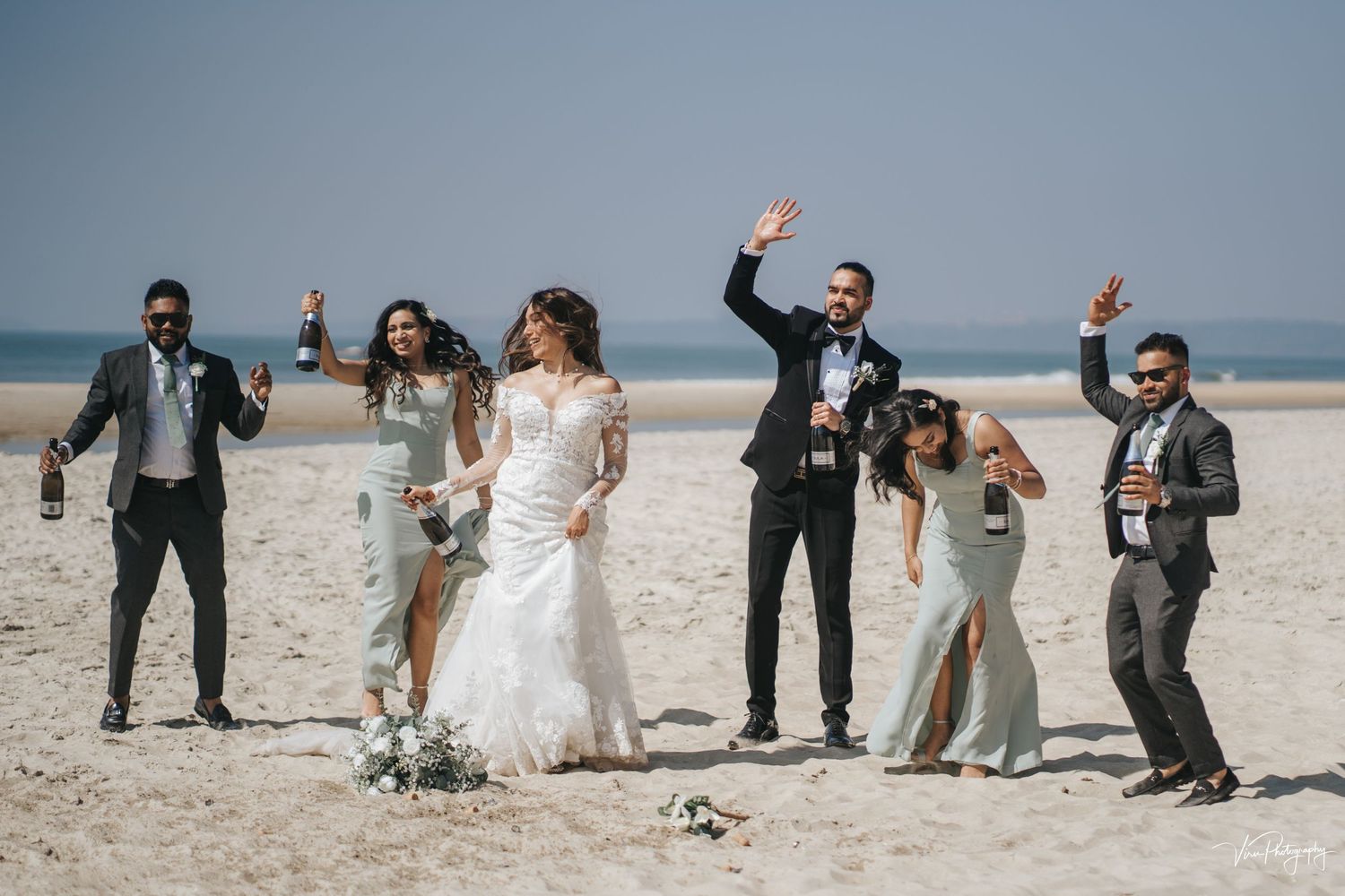 A joyful wedding party celebrates on a sunny beach, raising their arms in celebration against the ocean backdrop.