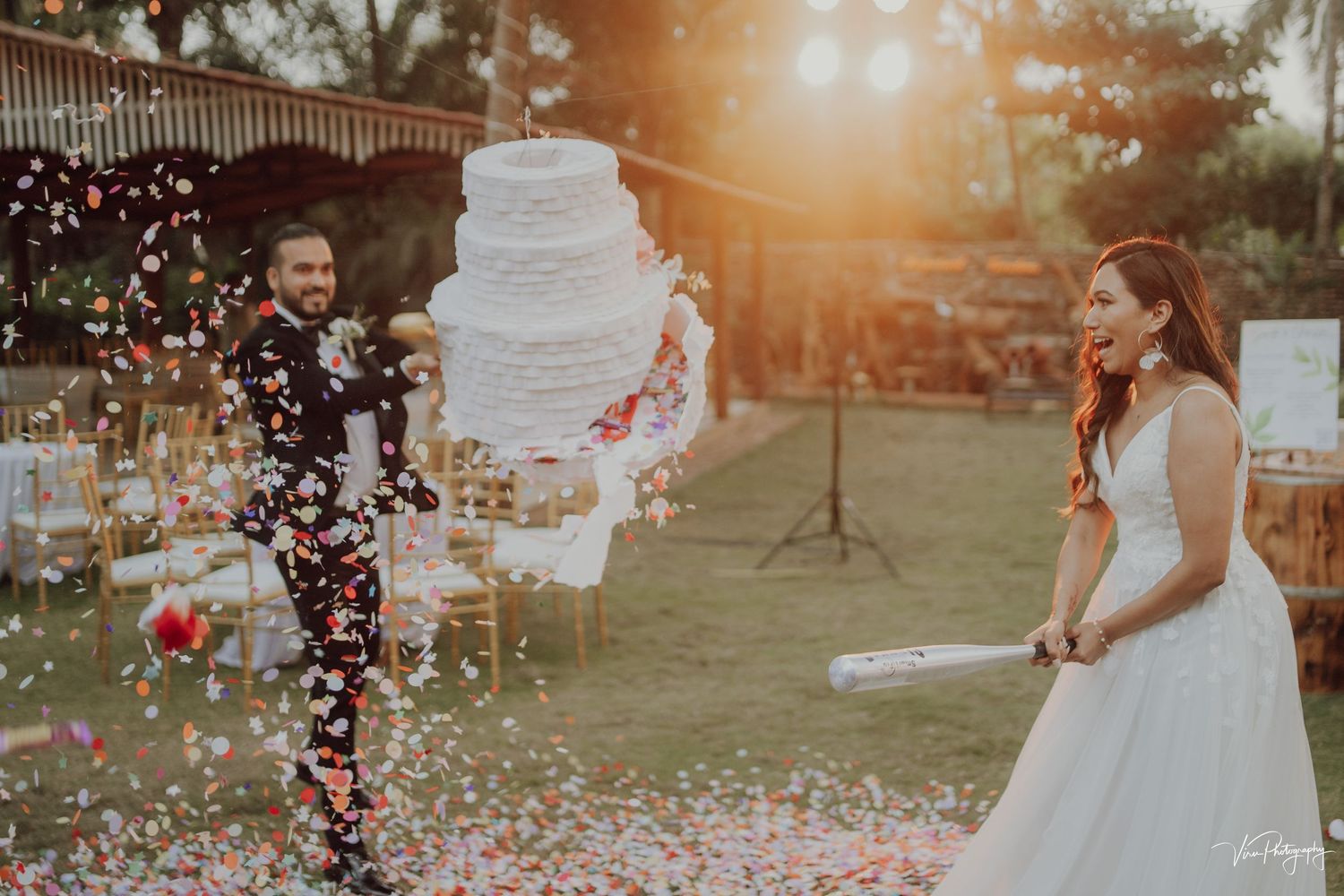 Wedding couple celebrates with confetti and a white tiered cake in outdoor garden setting at sunset.