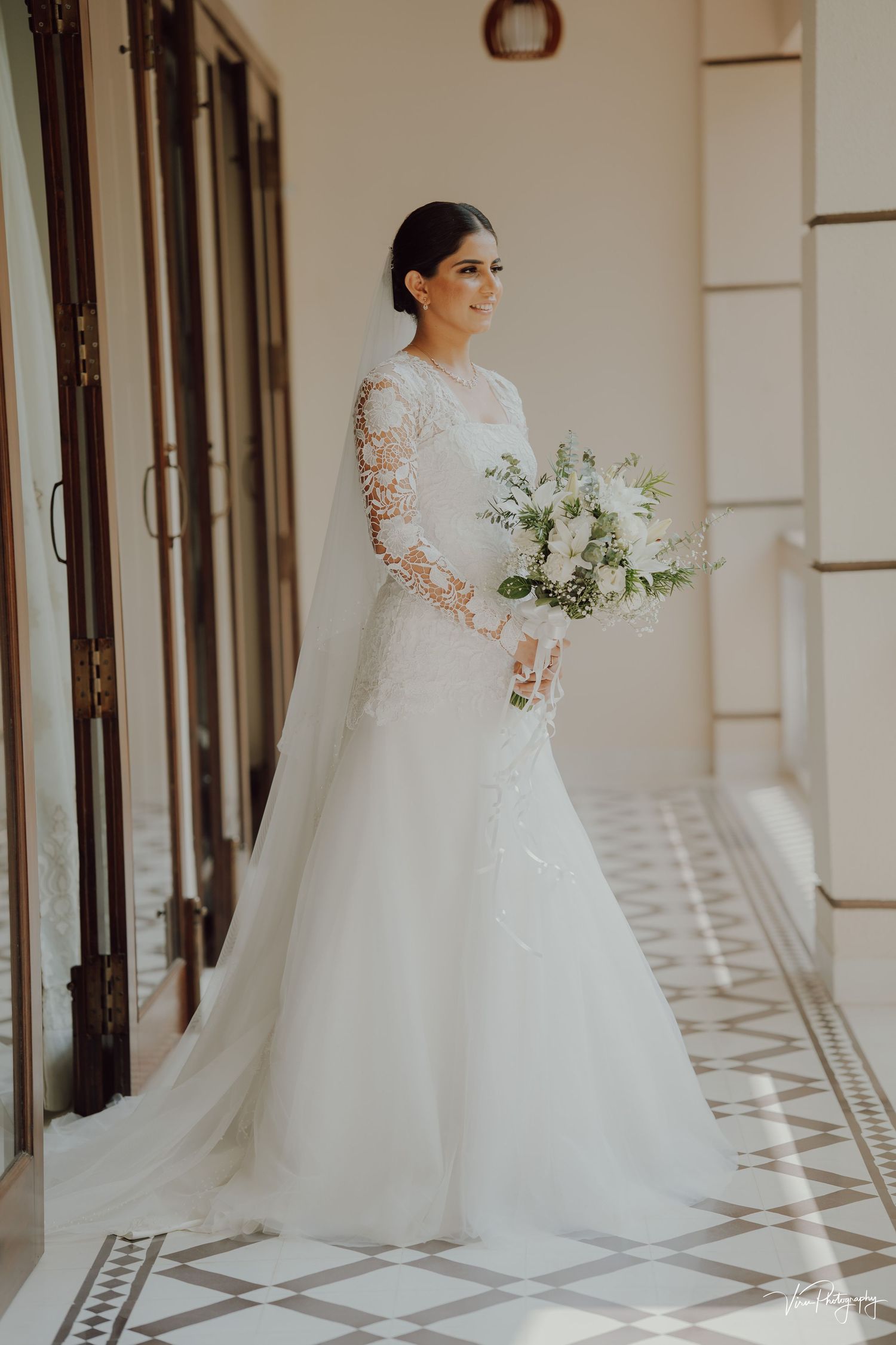 Elegant bride in long-sleeved lace gown with flowing train holds white floral bouquet in sunlit hallway with patterned floor.