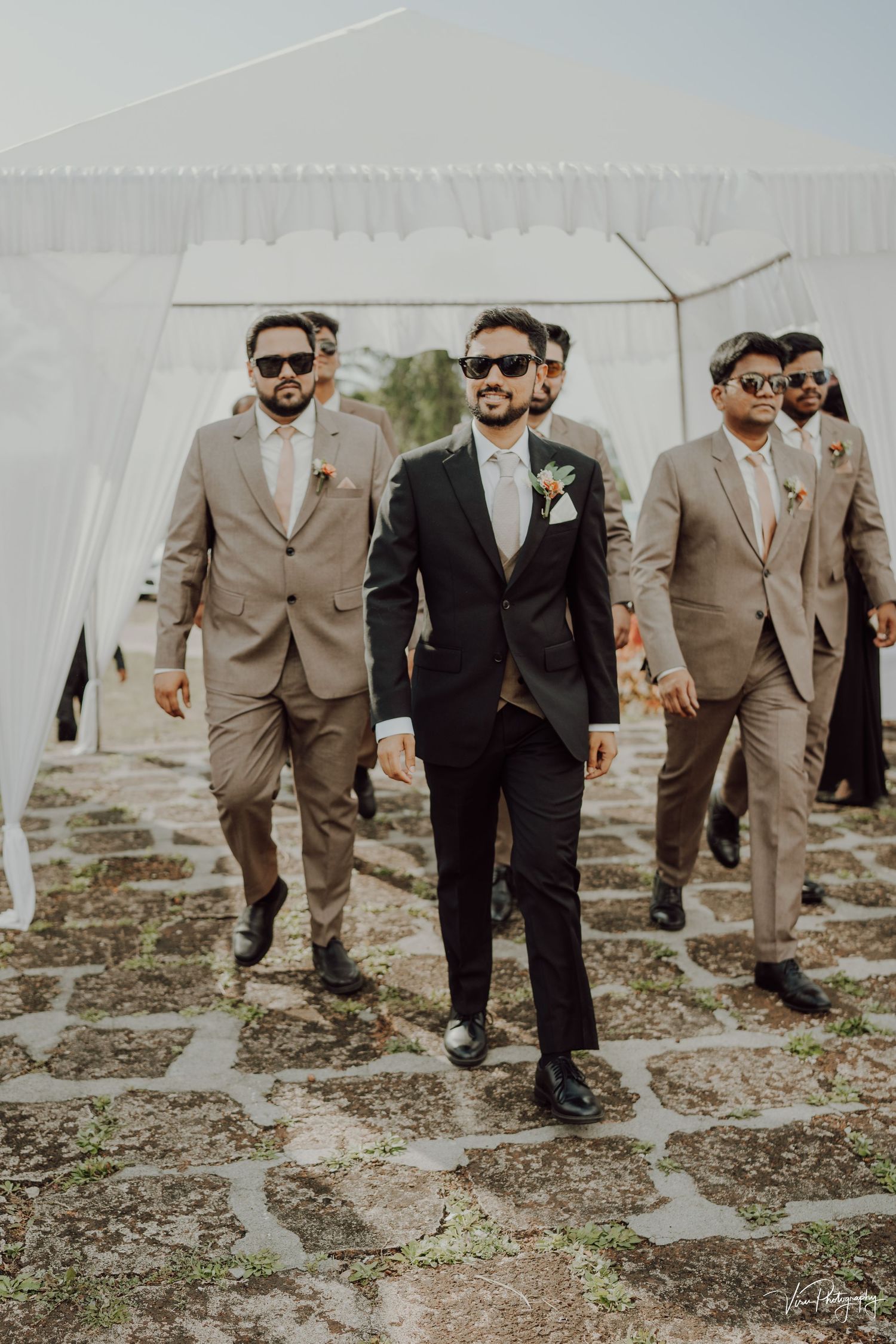 Groomsmen wearing sunglasses at outdoor wedding walking together in matching tan suits with one in black.