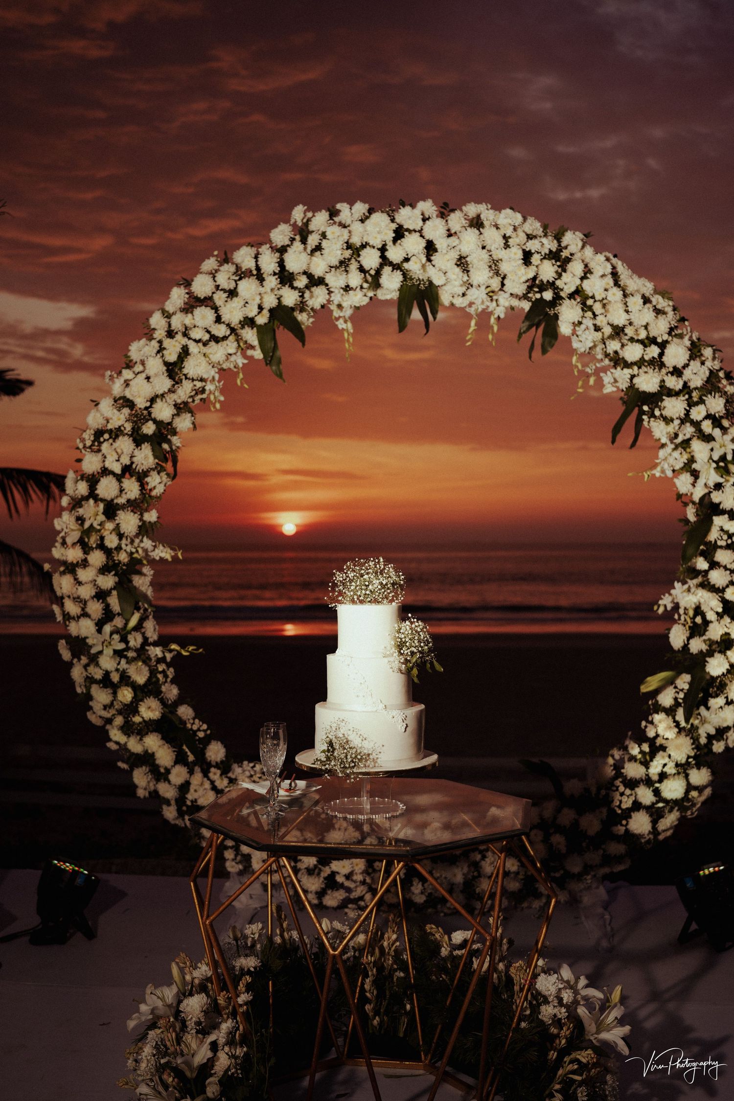 A white wedding cake displayed under a floral arch against a dramatic orange sunset over the ocean backdrop.