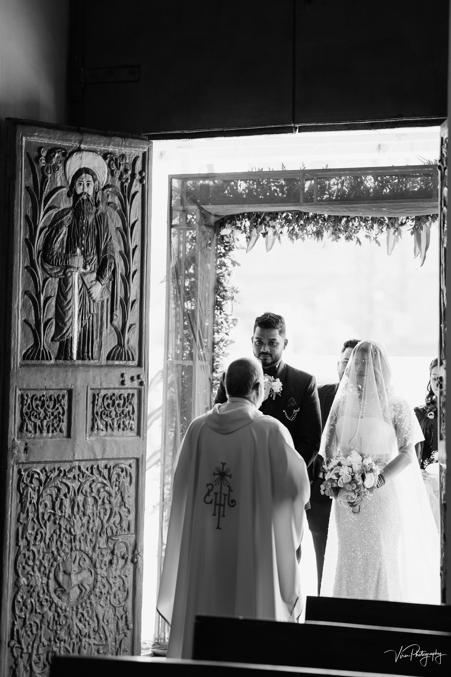 A black and white wedding ceremony photograph showing a decorated church doorway with an officiant performing the service.