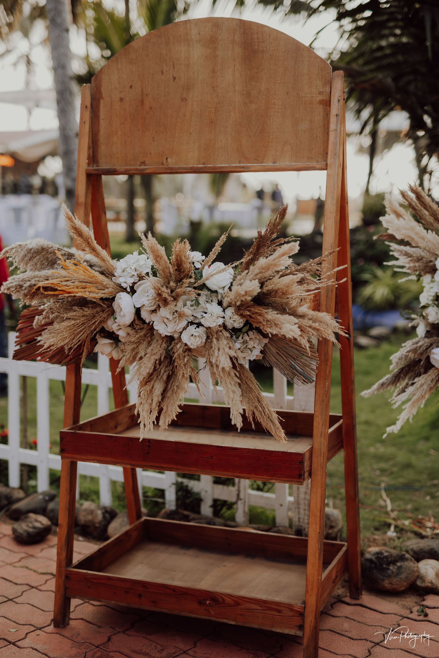 A rustic wooden display ladder decorated with dried pampas grass and floral arrangements for an outdoor wedding ceremony.
