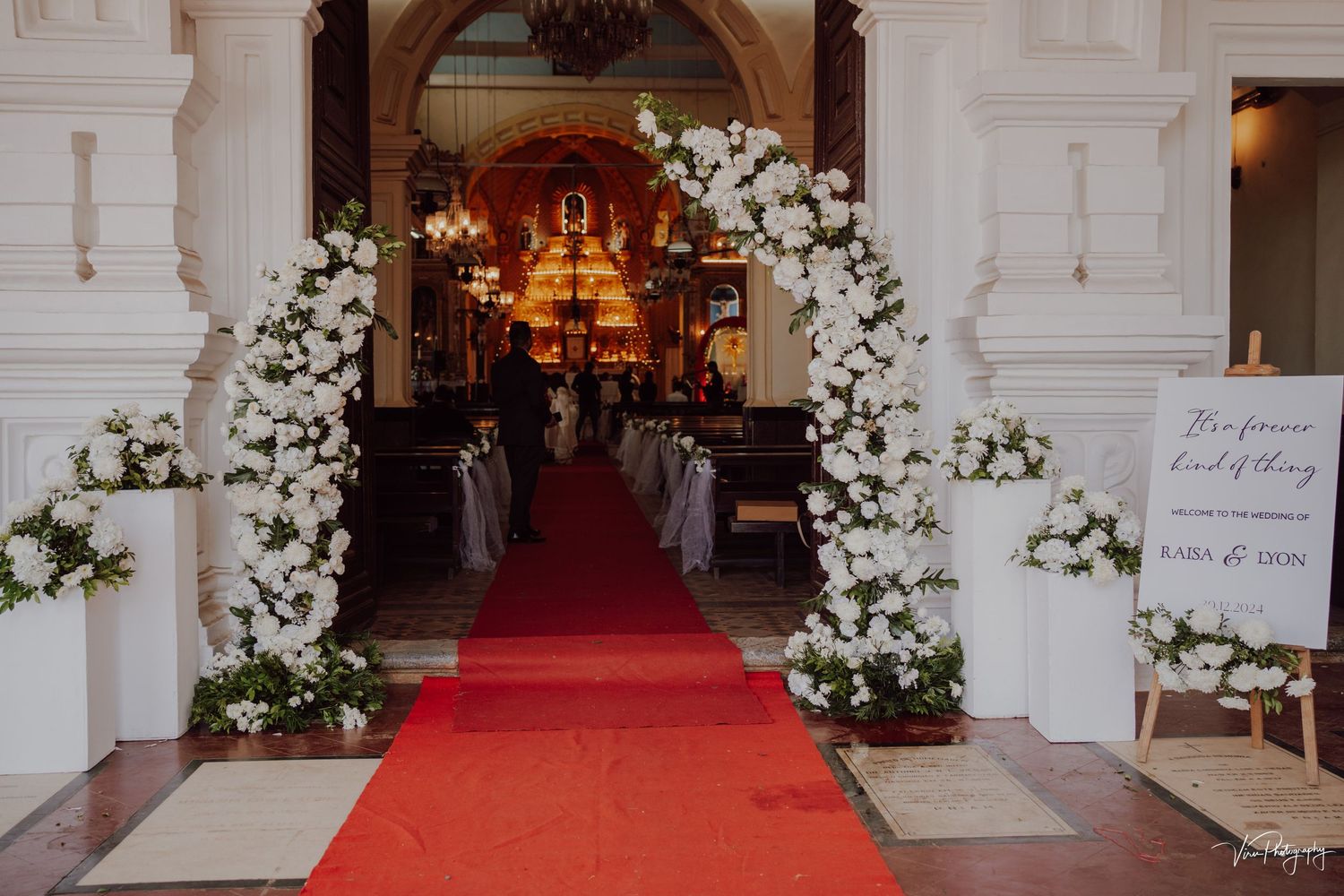 Elegant church entrance decorated with white floral arches and red carpet leading to illuminated altar.