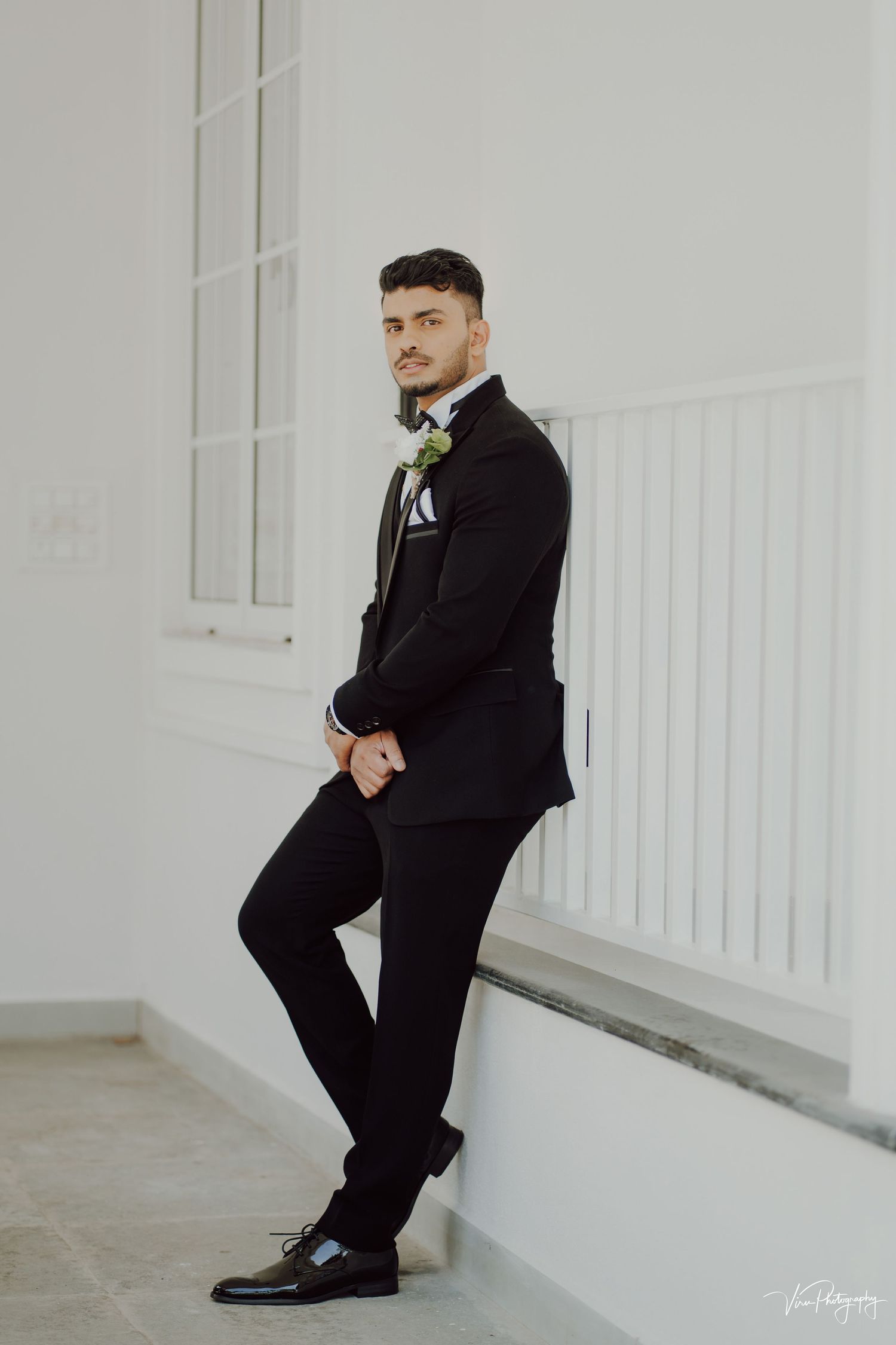 Someone in a classic black tuxedo with boutonniere leans against white wall in elegant wedding attire.