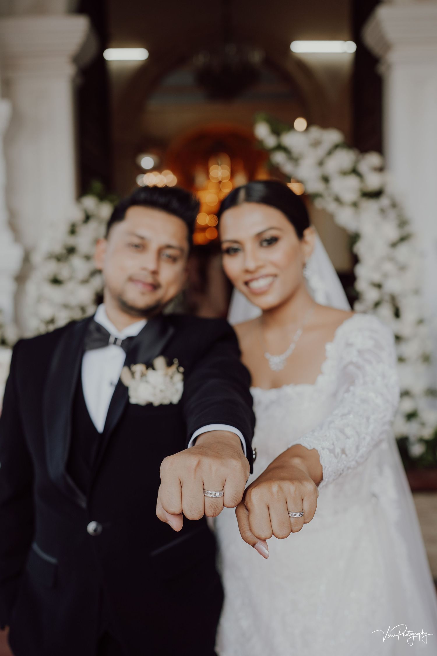 Newlywed couple showing off their wedding rings during an elegant ceremony with white floral decorations.