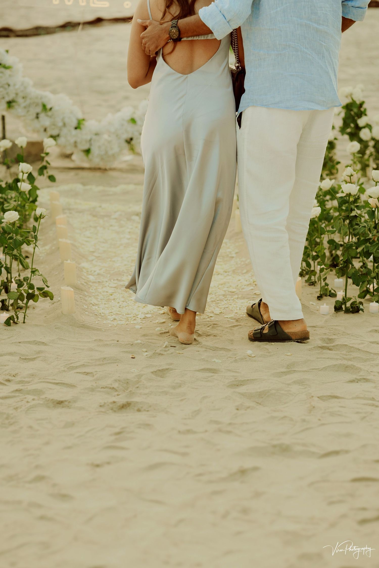 Couple walking barefoot on sandy beach with flowing dress and casual attire on a sunny day.