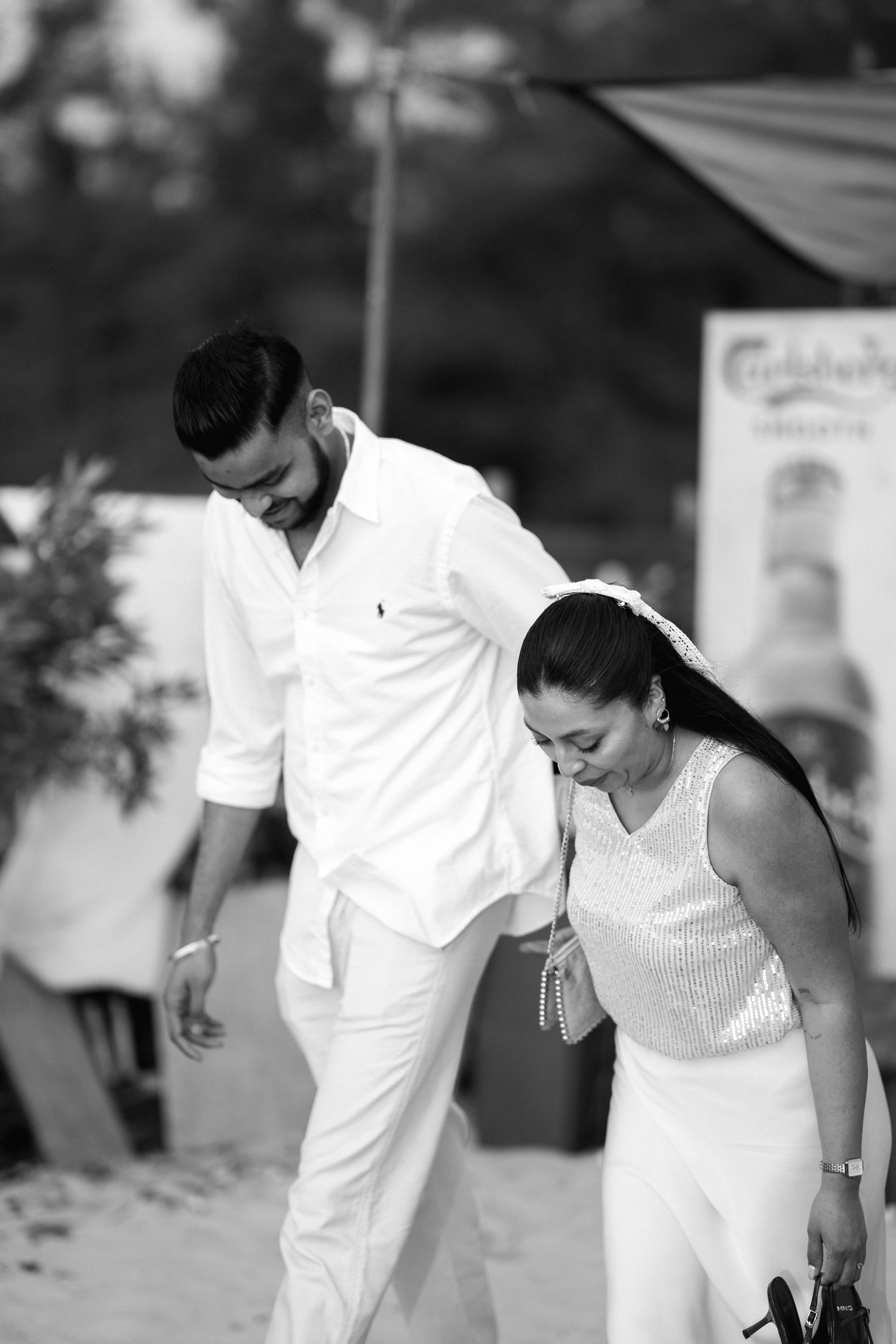 Black and white photo of a couple walking on a beach, man in white shirt and pants, woman in sequined top.