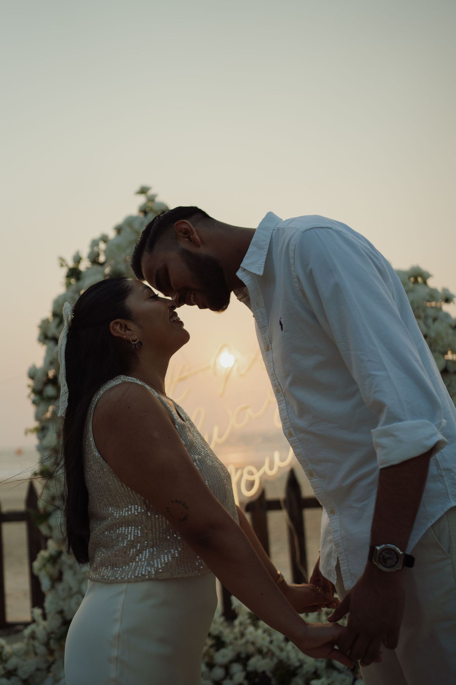 Romantic couple holding hands at sunset near a floral arch with neon lights during an outdoor proposal.
