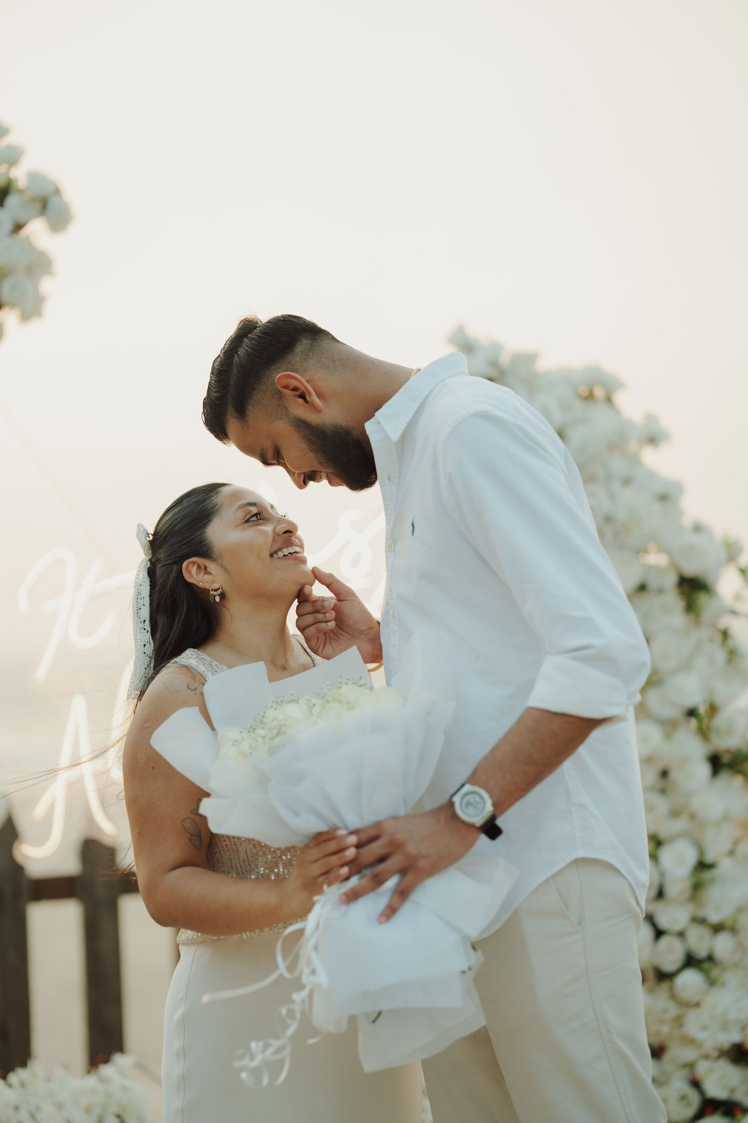 A couple in white attire shares a romantic moment outdoors, with the bride holding a white bouquet near a floral arch.