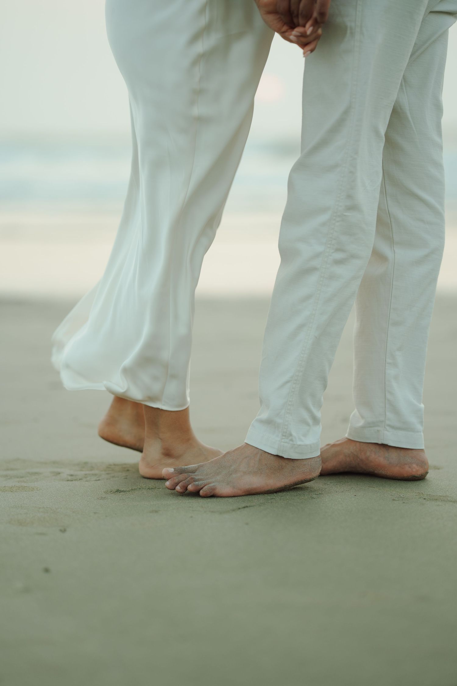 Barefoot couple in white pants standing on sandy beach, holding hands romantically near ocean waves.