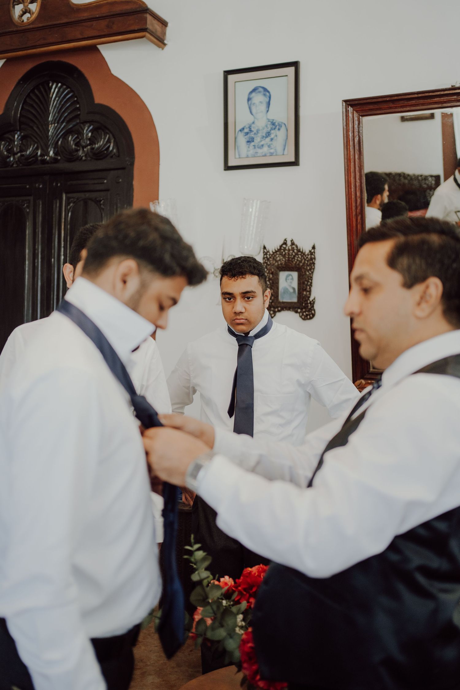 Three groomsmen in white shirts and ties help each other get ready for a formal event.