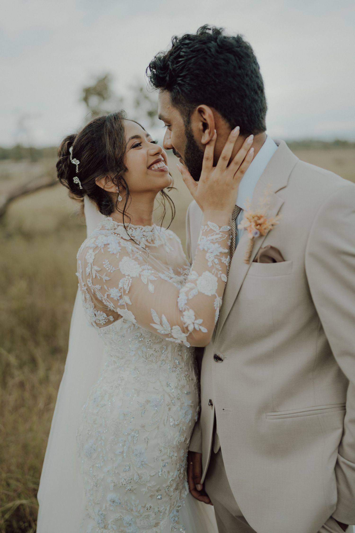 A joyful bride in an embroidered lace gown touches her groom's face as he smiles, wearing a beige suit with an orange boutonniere.