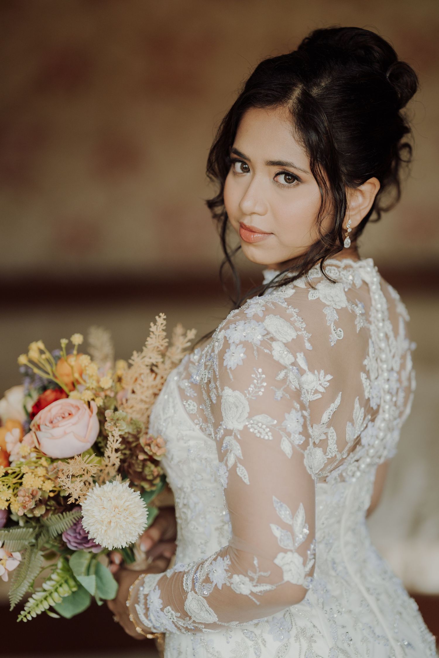 Bride in a lace long-sleeve wedding gown with an elegant updo holds a colorful bouquet and looks over her shoulder.