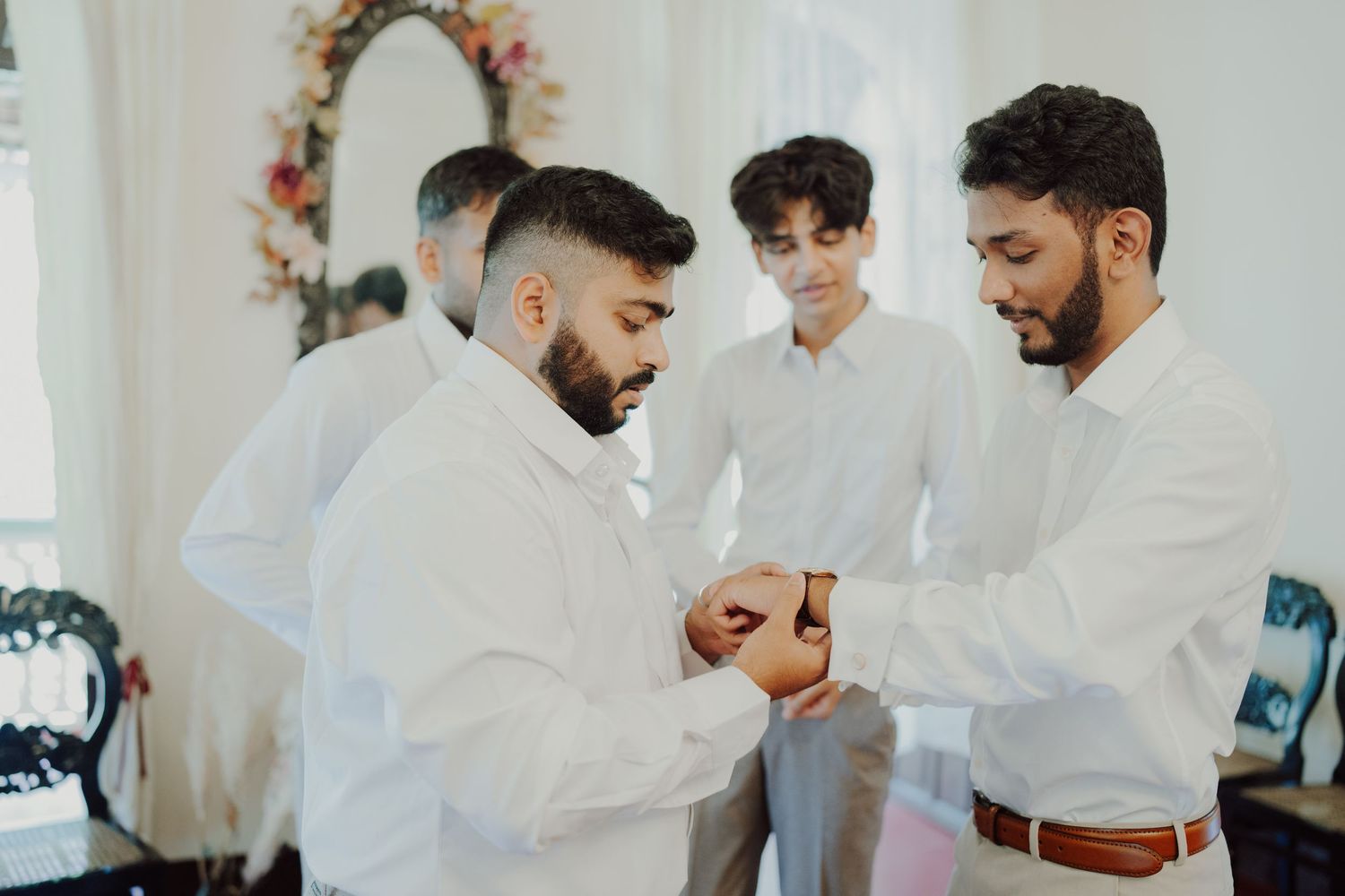 Groomsmen in white dress shirts help one another get ready, adjusting a watch on the wrist in a bright room before a wedding ceremony.