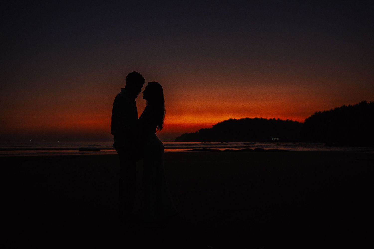 Silhouette of a romantic couple embracing on a beach at sunset with vibrant red and orange sky.