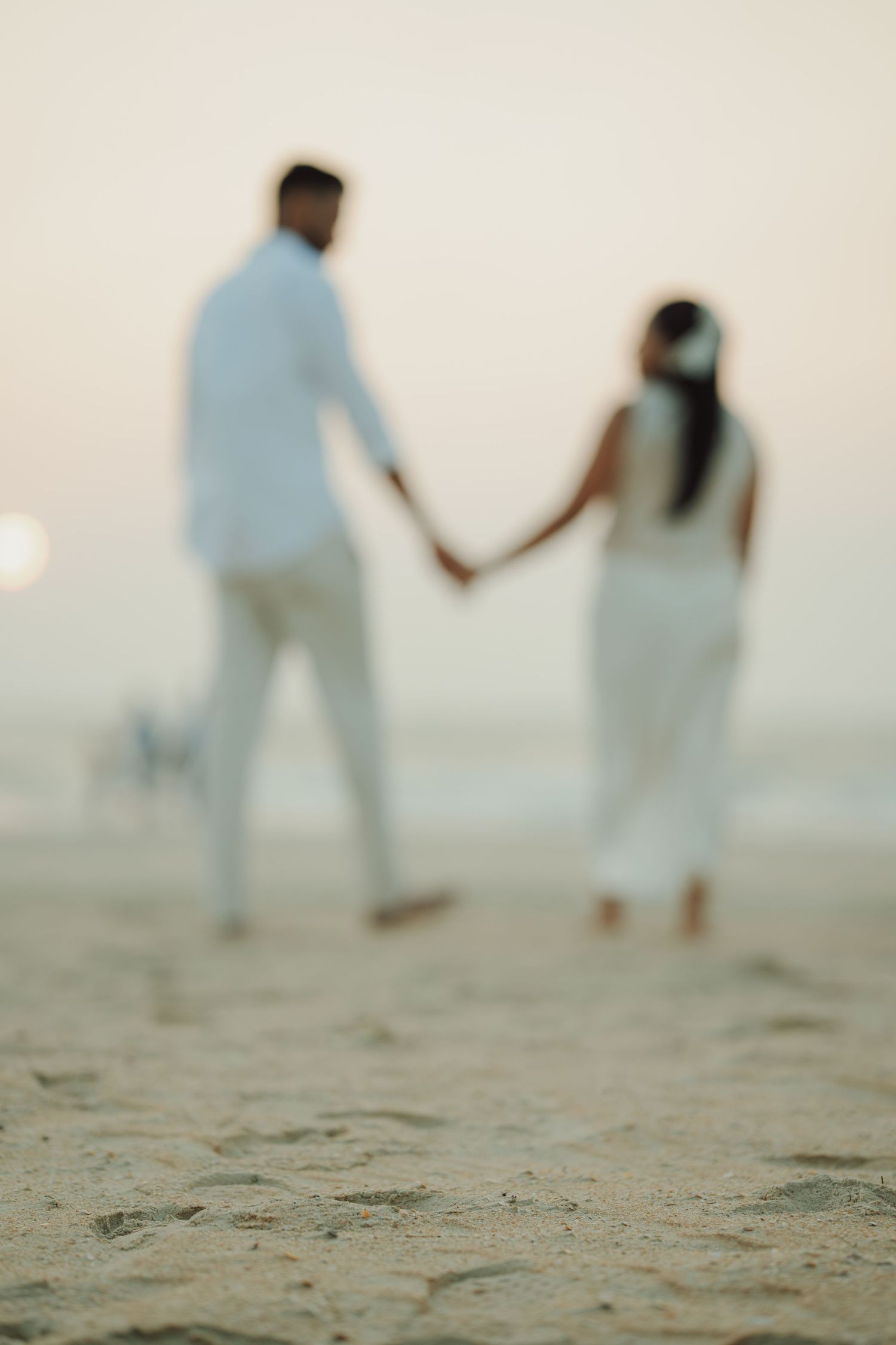 Blurred couple in white holding hands on a sandy beach at sunset, romantic seaside wedding scene.