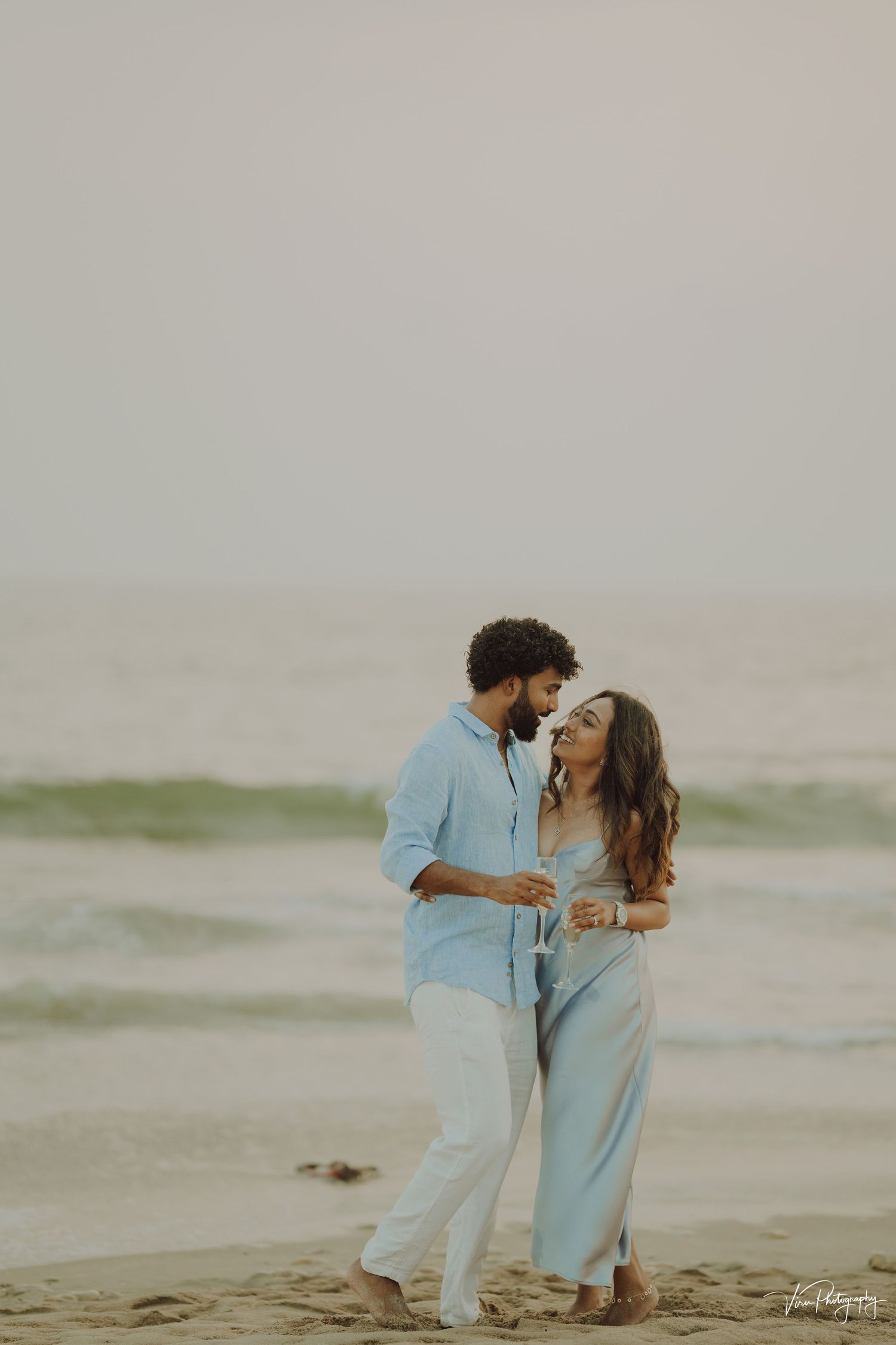 Couple in matching light blue outfits share a romantic moment while walking along a misty beach at sunset.