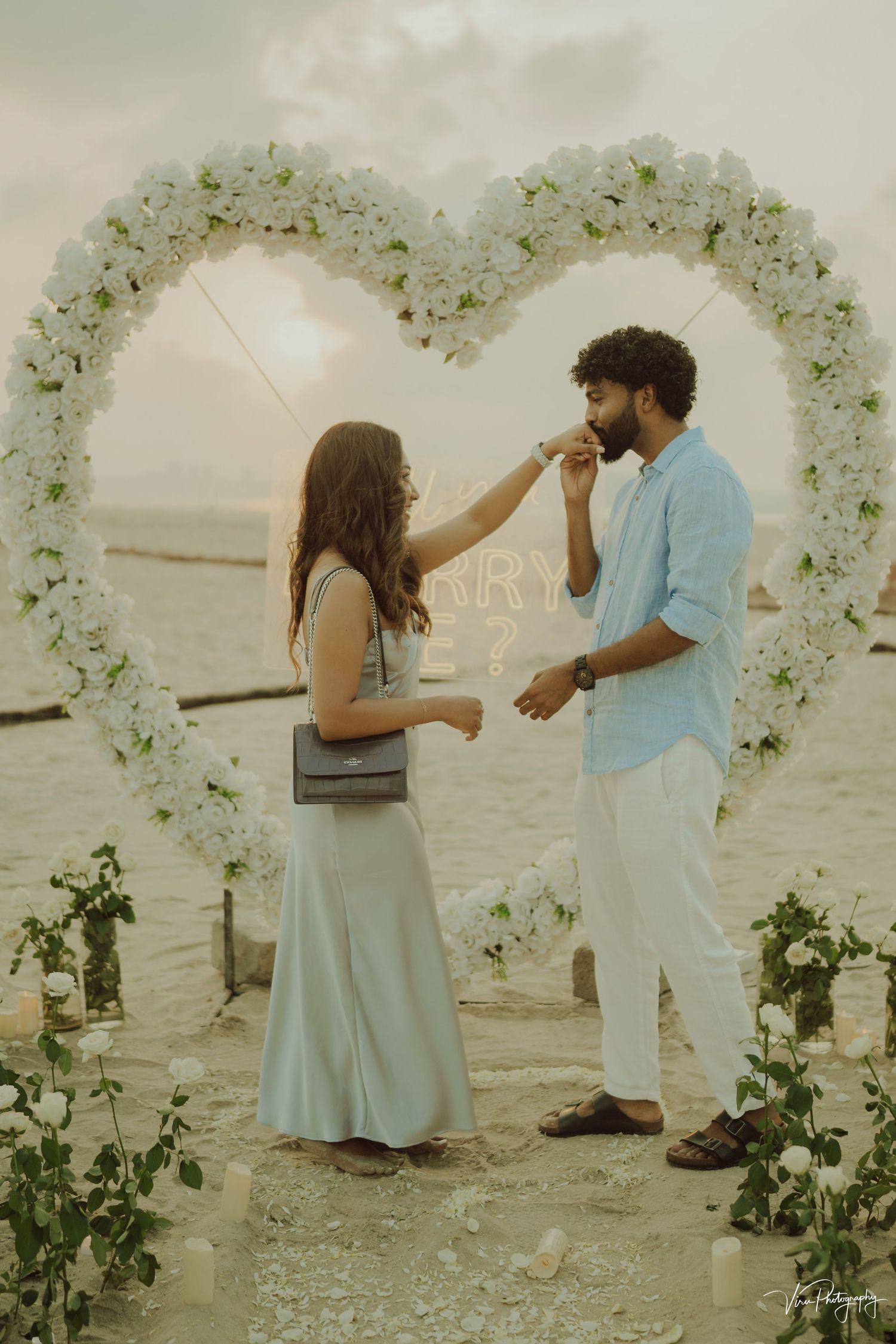 A romantic beach proposal scene with two people standing under a white floral heart-shaped arch during golden hour.