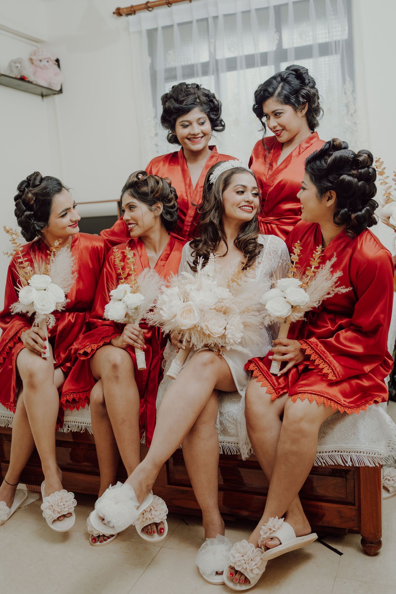 Bridal party in matching red silk robes and fuzzy slippers sharing a candid laughing moment together on wedding day.