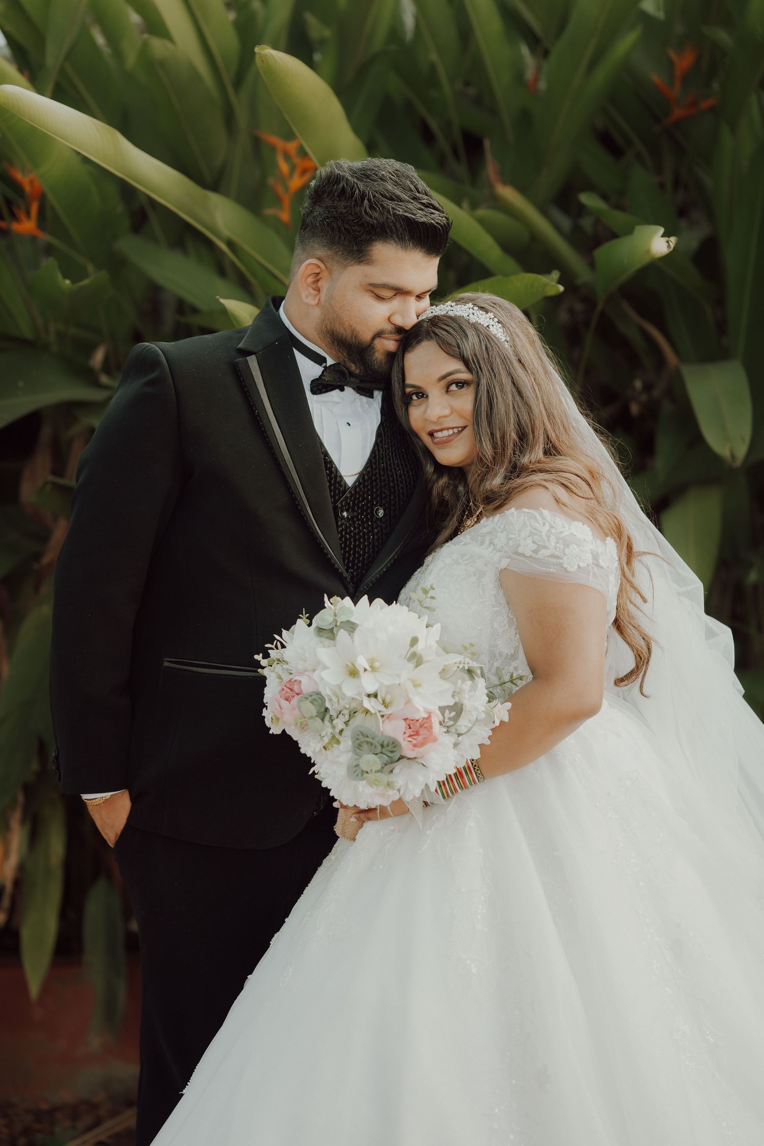 A bride in a white ballgown holds a pink and white bouquet while posing with her groom in a black tuxedo against lush foliage.
