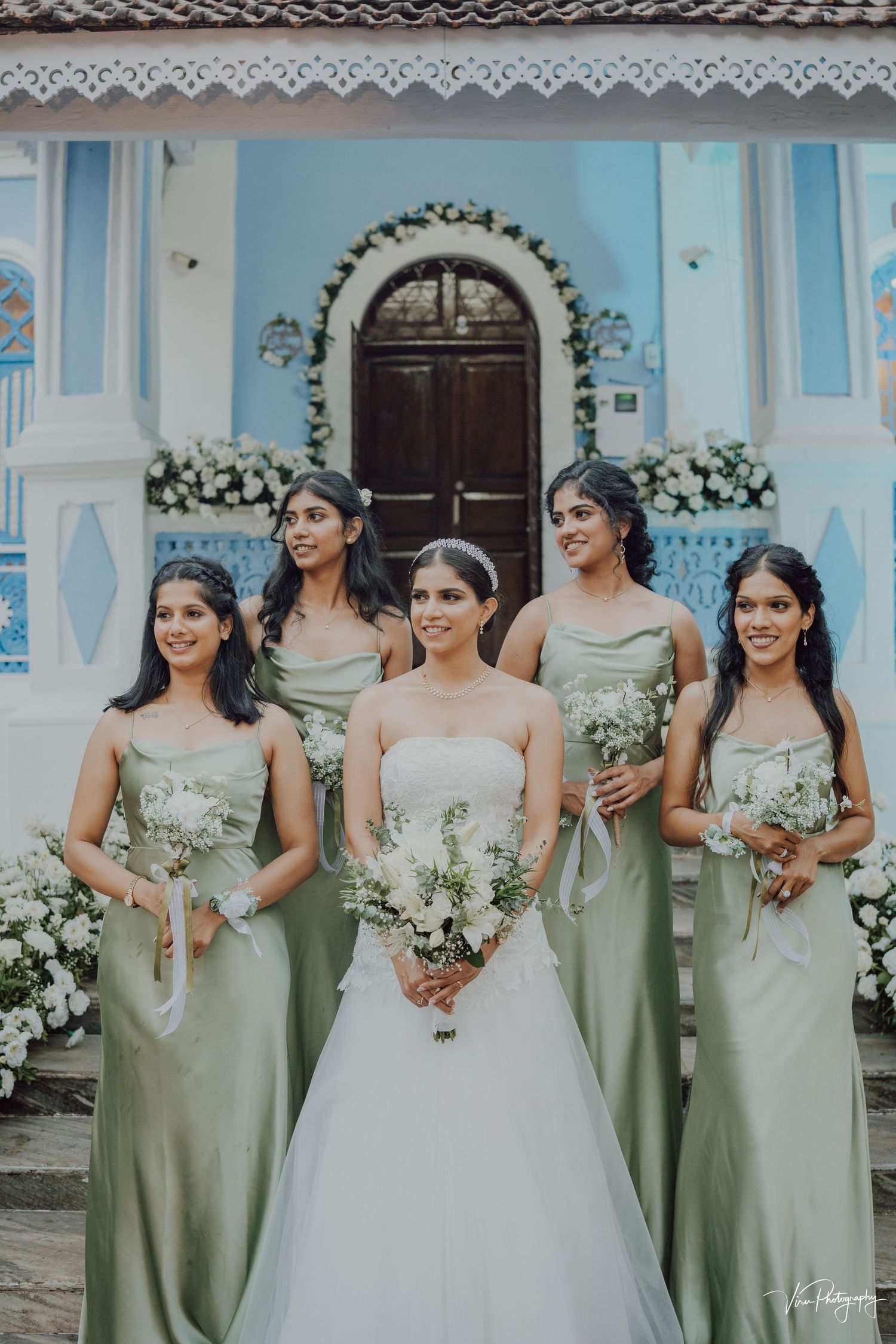 A bridal party in mint green dresses poses in front of a blue church with ornate floral archway.