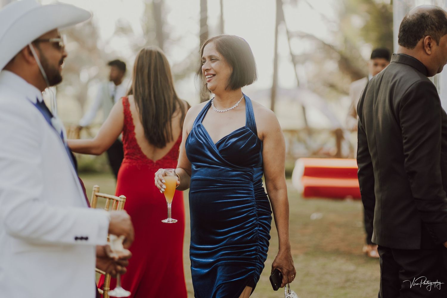 Guests mingle at an elegant outdoor event, with someone wearing a blue silk dress holding a cocktail glass.