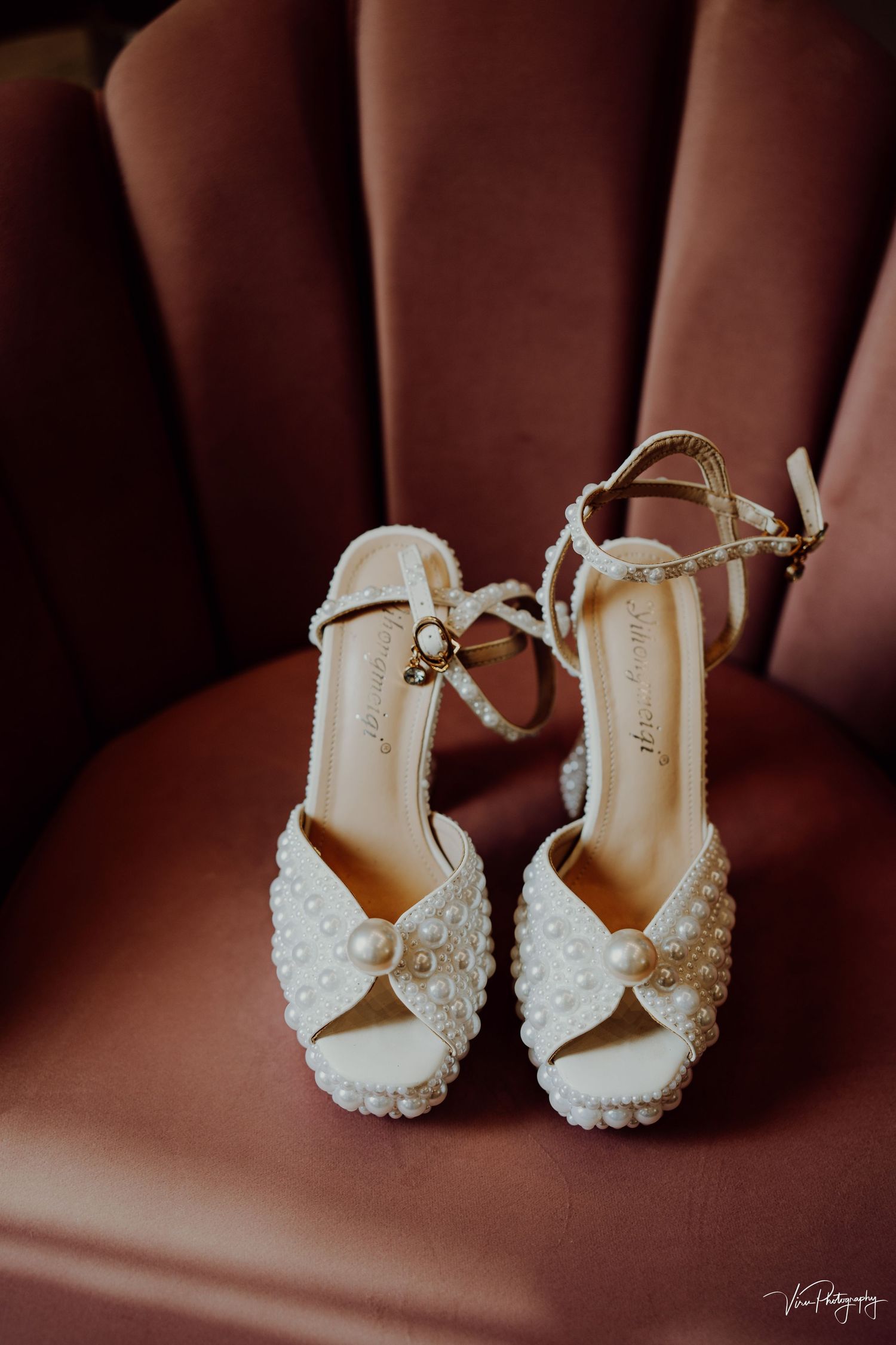 Elegant white beaded wedding sandals with pearl embellishments and ankle straps displayed on brown leather chair.