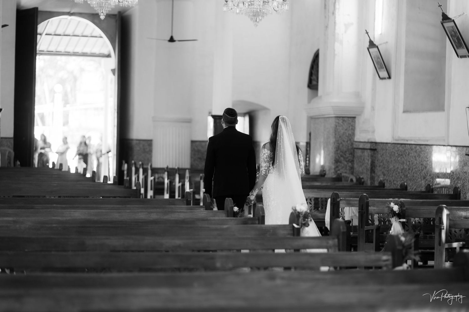 A black and white photograph of a couple walking through church pews towards an arched doorway in the distance.