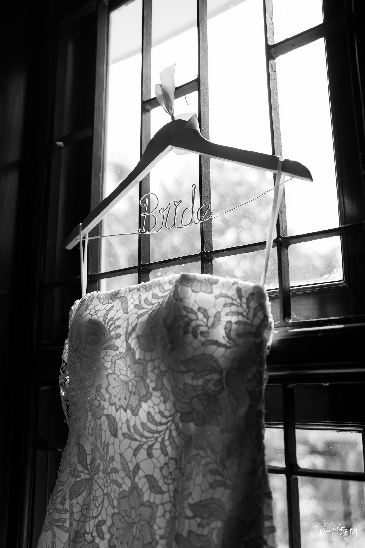 A lace wedding dress hangs on a wooden hanger near a window in black and white photography.